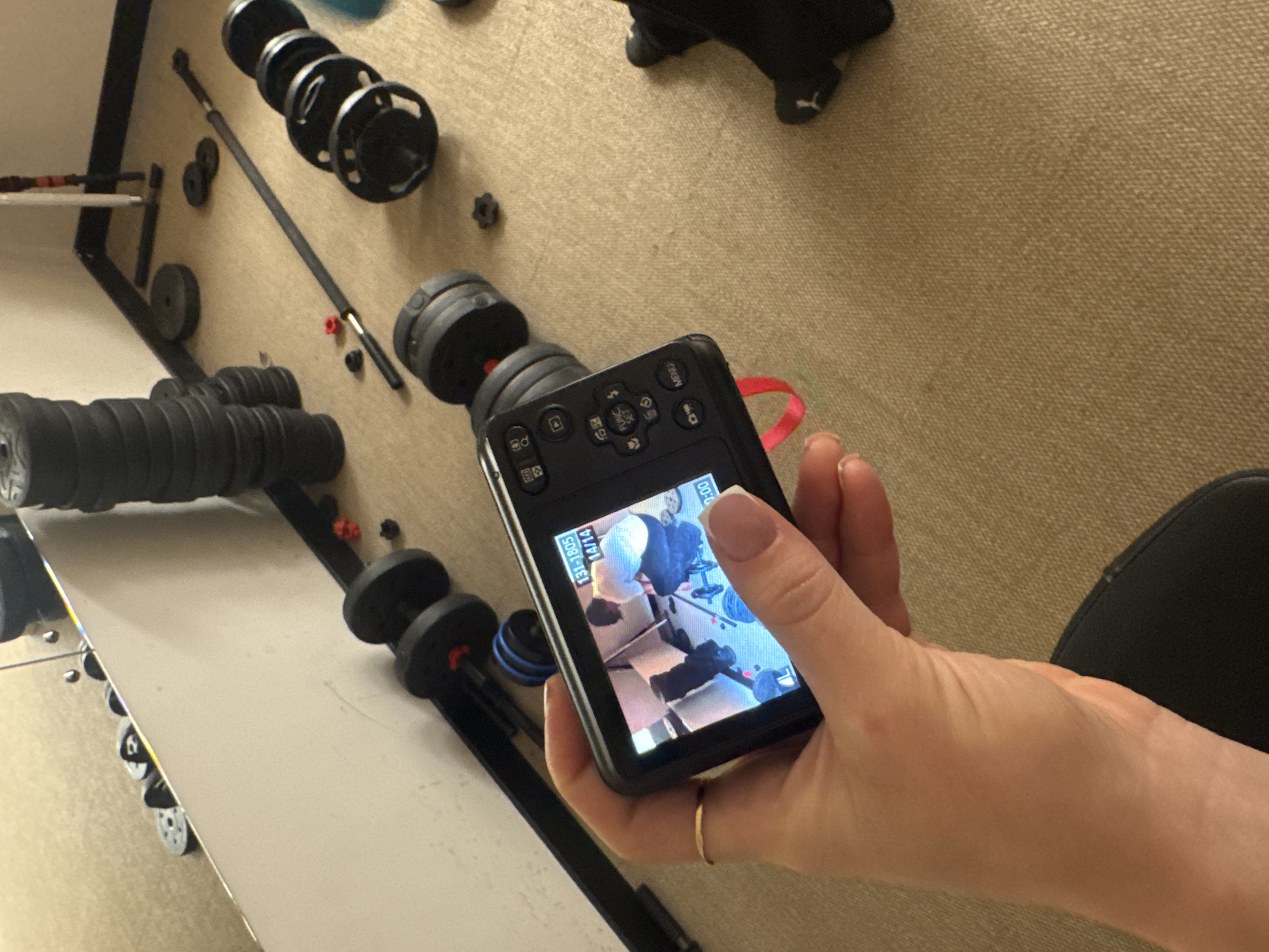 A digital camera is held in a person's hand, displaying an image of weightlifting equipment. The surrounding environment includes a beige textured surface, likely carpet, and various pieces of weightlifting equipment such as dumbbells and weight plates. The location, Khimki, Russia is not directly discernible from the image.