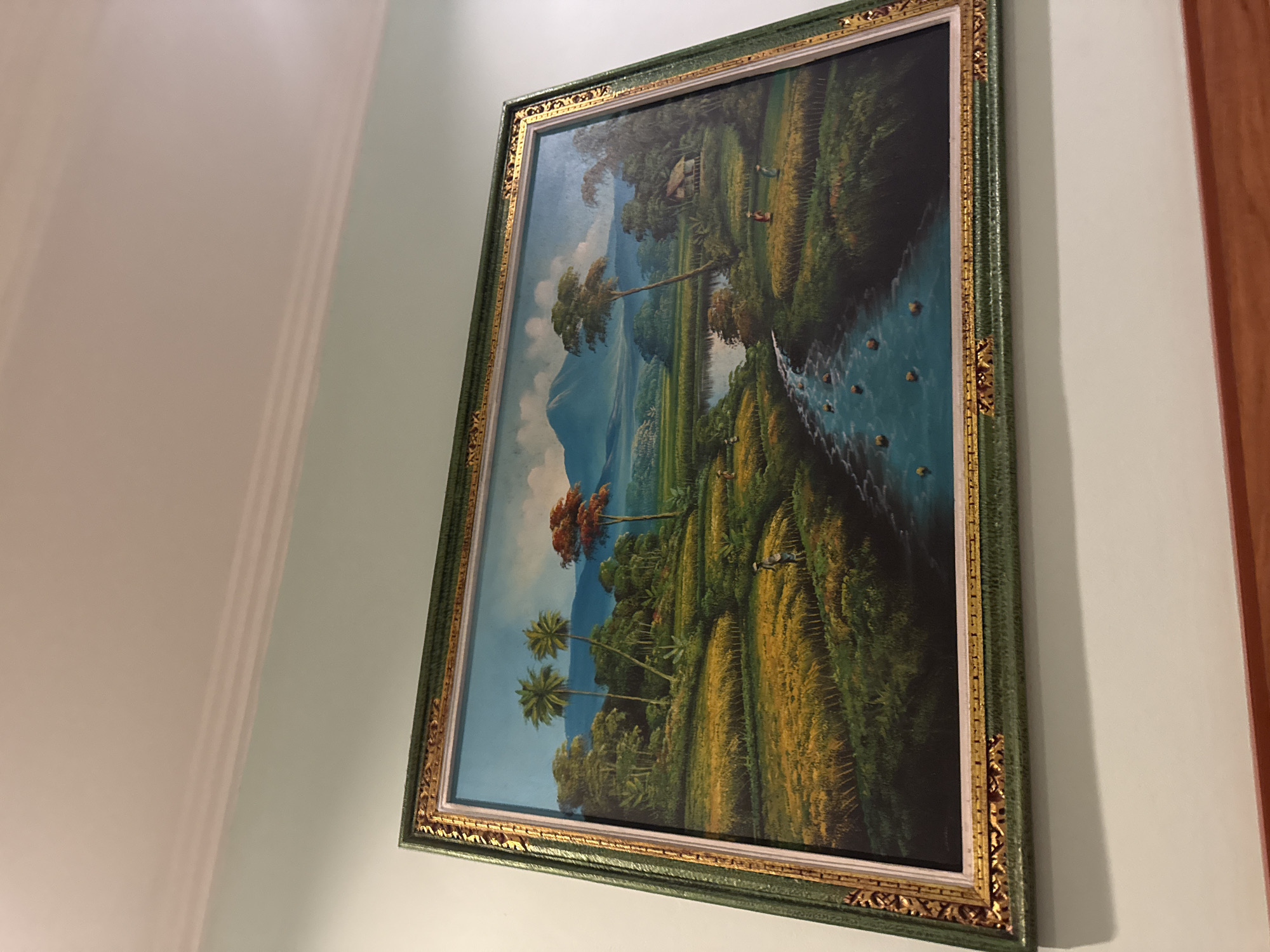 A framed painting depicting a vibrant, panoramic landscape hangs on a light-colored wall. The painting, oriented vertically, showcases a lush tropical scene consistent with the Ubud District in Indonesia. In the background, majestic blue mountains rise against a clear sky dotted with a few white clouds.
The middle and foreground are dominated by extensive, golden-yellow rice paddies, terraced across the undulating terrain, suggesting a harvest-ready crop. Numerous tall palm trees and other dense green foliage are scattered throughout the landscape, particularly along the banks of a winding, blue-green river or stream that flows from the upper right to the lower left of the canvas. Small, dark shapes are visible within the water, possibly indicating rocks or small boats.
Several small figures, presumably people, are depicted working within the rice fields, adding a sense of human activity to the serene natural setting. A traditional-looking hut is nestled among trees in the upper left portion of the painting. The painting is encased in an elaborate frame, featuring a deep green border with intricate, gold-colored decorative patterns along its inner and outer edges. The scene evokes a tranquil depiction of traditional agricultural life in a beautiful tropical environment during daytime. No text is visible in the image.