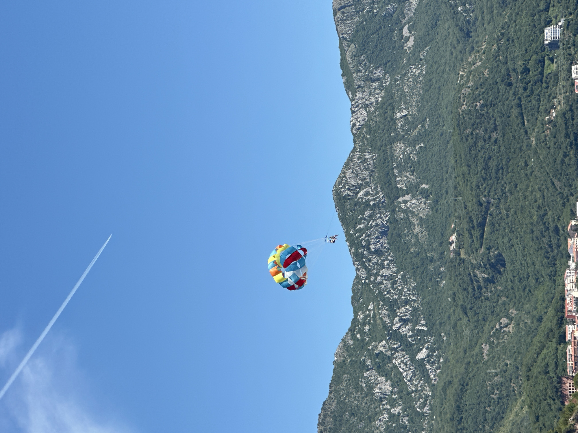 A bright, colorful parasail with two people attached flies through a clear blue sky above a lush green mountainous landscape. A faint white contrail from an airplane stretches across the upper left quadrant of the sky. The mountainside is densely covered in trees with patches of rocky outcrops. In the distance, a coastal town with red-tiled roofs is visible at the base of the mountains. The image captures a sunny day with excellent visibility, suggesting a popular tourist destination.