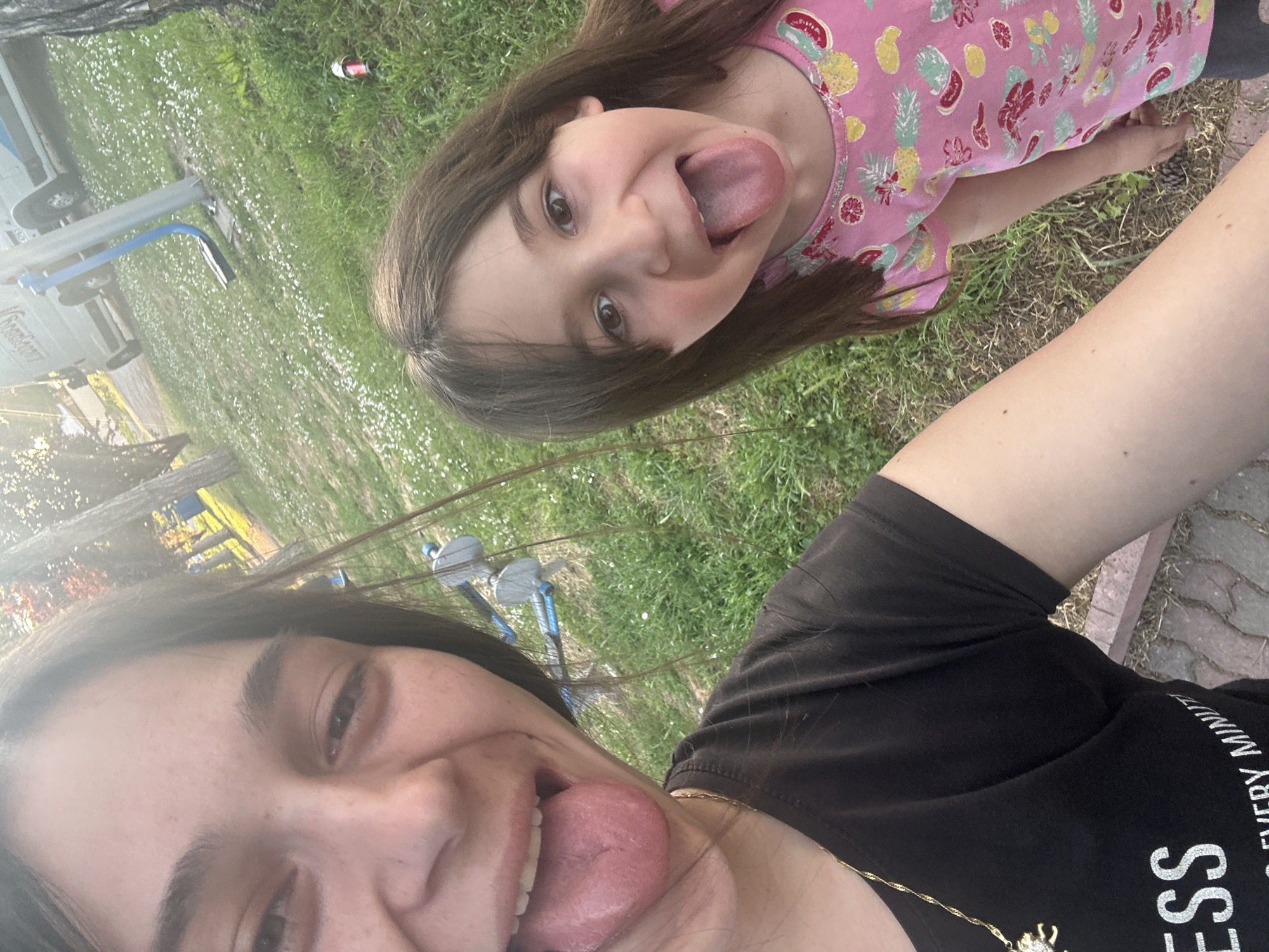 Two individuals, a child and an adult, are captured in a close-up, tilted photograph taken outdoors. Both individuals are sticking out their tongues. The child wears a pink top with a fruit pattern, while the adult wears a black t-shirt and a gold necklace. The background shows a grassy area with small white flowers, a playground structure, and a glimpse of a paved area. A discarded bottle is visible on the grass. The location may be within the city of Resen, Bulgaria.