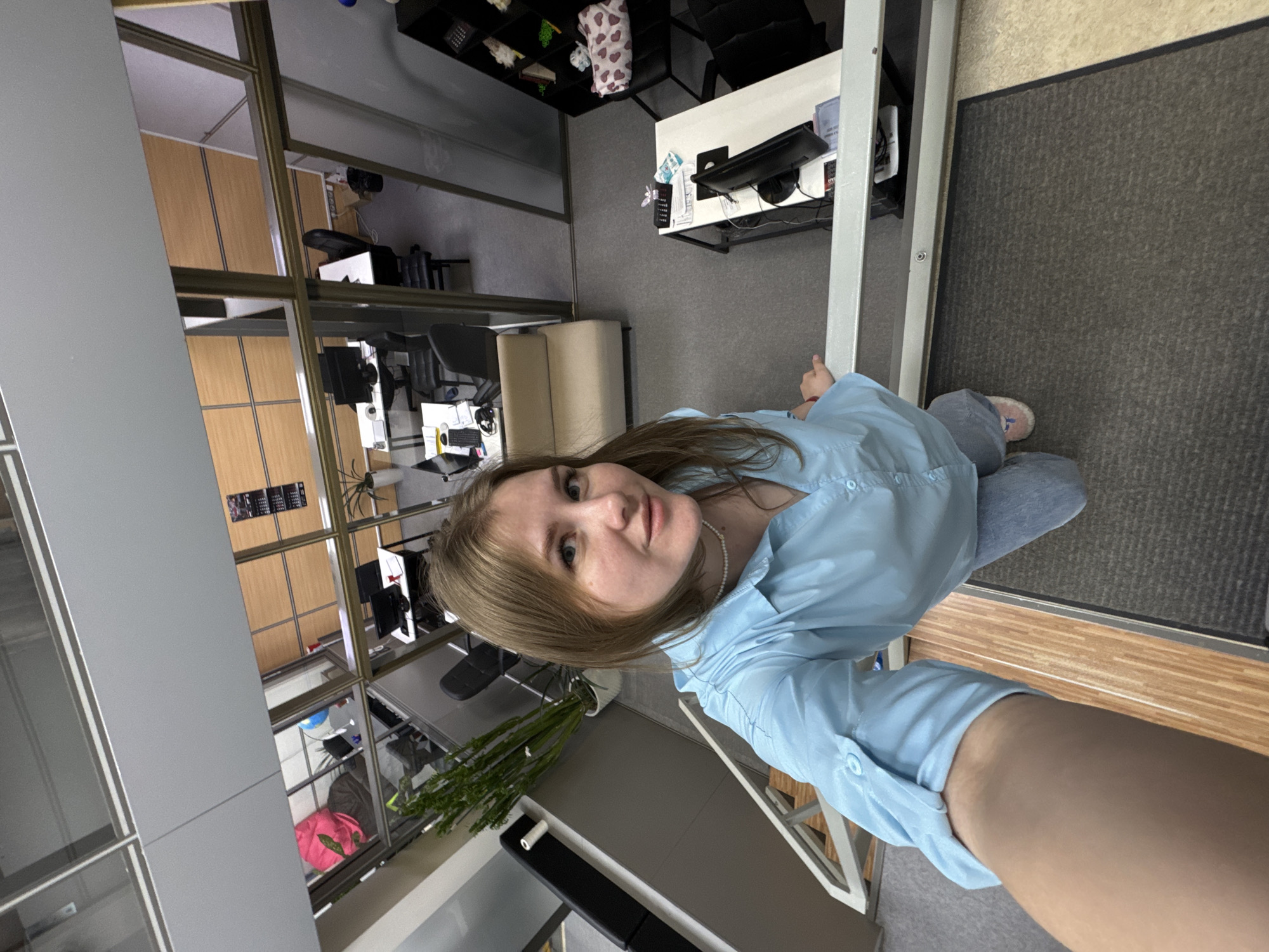 A young woman is depicted in a selfie taken inside what appears to be an office environment. She is fair-skinned with light brown hair and is wearing a light blue long-sleeved shirt. The setting features office desks and chairs, suggesting a modern office space, and the photograph's perspective indicates she may be in a multi-story building looking down at the office area. The location is identified as the city of Perm, Russia. Other details include flooring, a partition wall, and office plants.