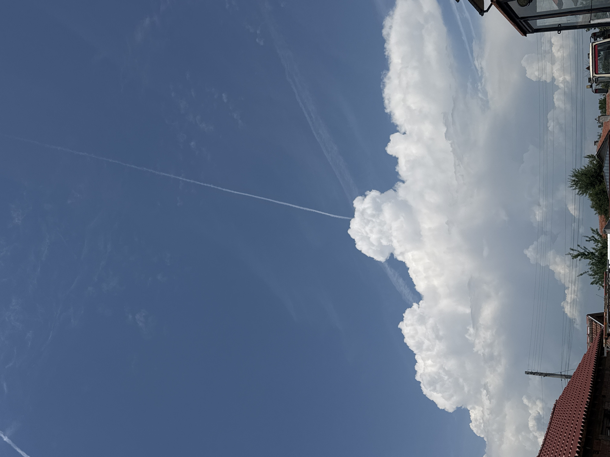 This image, recorded in Sarnevo, Bulgaria, presents a clear blue sky largely occupied by substantial cloud formations. A distinct, linear white contrail traverses the upper left quadrant, extending diagonally downwards towards the image center, where it intersects a massive, vertically developed cumuliform cloud. A second, broader and more dispersed contrail-like streak is observable horizontally across the mid-right sky. The lower two-thirds of the frame are dominated by this expansive, brilliantly white cumulus or cumulonimbus cloud, characterized by rounded, cauliflower-like tops. Beneath the cloud mass, the upper portions of human-made structures and vegetation are visible. In the lower left, a residential structure with a red, ridged terracotta-tiled roof and partial brick wall is present. A utility pole supporting multiple parallel power lines stands near the center-left, with the lines extending horizontally across the mid-ground. Further right, additional red-tiled rooftops are partially obscured by green trees and shrubs. The lower right corner reveals part of a building featuring prominent glass panels and a segment of a white and red vehicle, consistent with a tractor. No human figures are depicted.