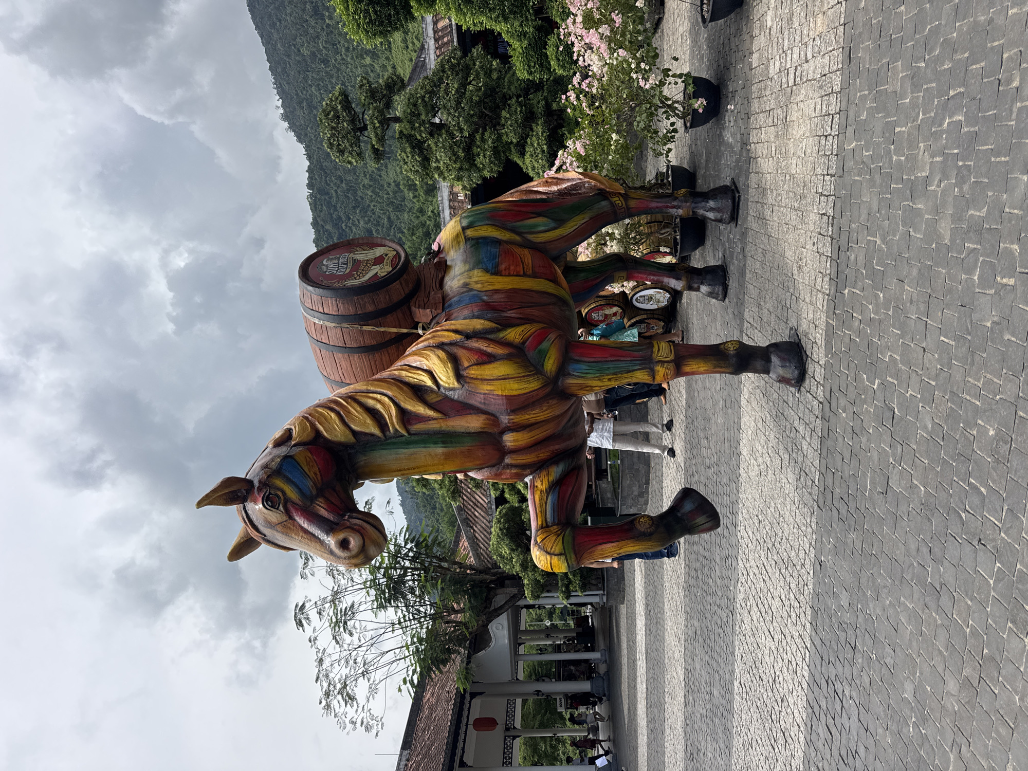 A brightly colored, horse-shaped statue with a barrel on its back is on display on a grey-bricked plaza in Đà Nẵng, Vietnam. The horse statue is painted with swirling patterns of red, yellow, blue, and brown. The barrel on its back is brown with red details. People can be seen walking in the background and sitting in what appears to be a temple. The sky is cloudy, and the backdrop contains lush green mountains.