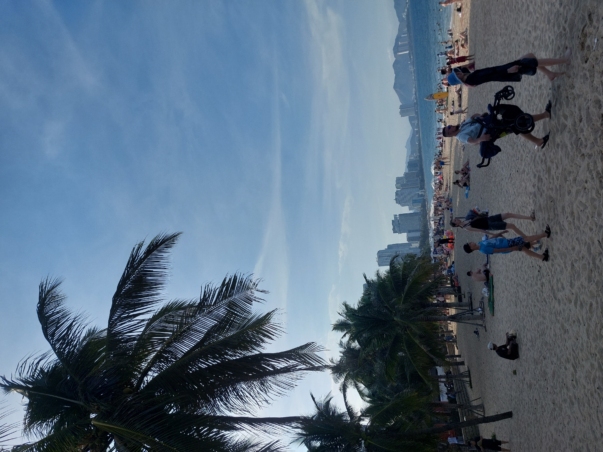 This is a photograph taken from a beach in Nha Trang, Vietnam, looking out towards the ocean. In the foreground, there is a sandy beach with several people scattered about. A man is pushing a stroller, and a woman is walking beside him. A young boy in a blue shirt and shorts is walking in front of them. In the middle ground, more people are relaxing on the sand and swimming in the ocean. There are palm trees lining the edge of the beach. In the background, there are tall buildings and mountains visible. The sky is blue with some scattered clouds.