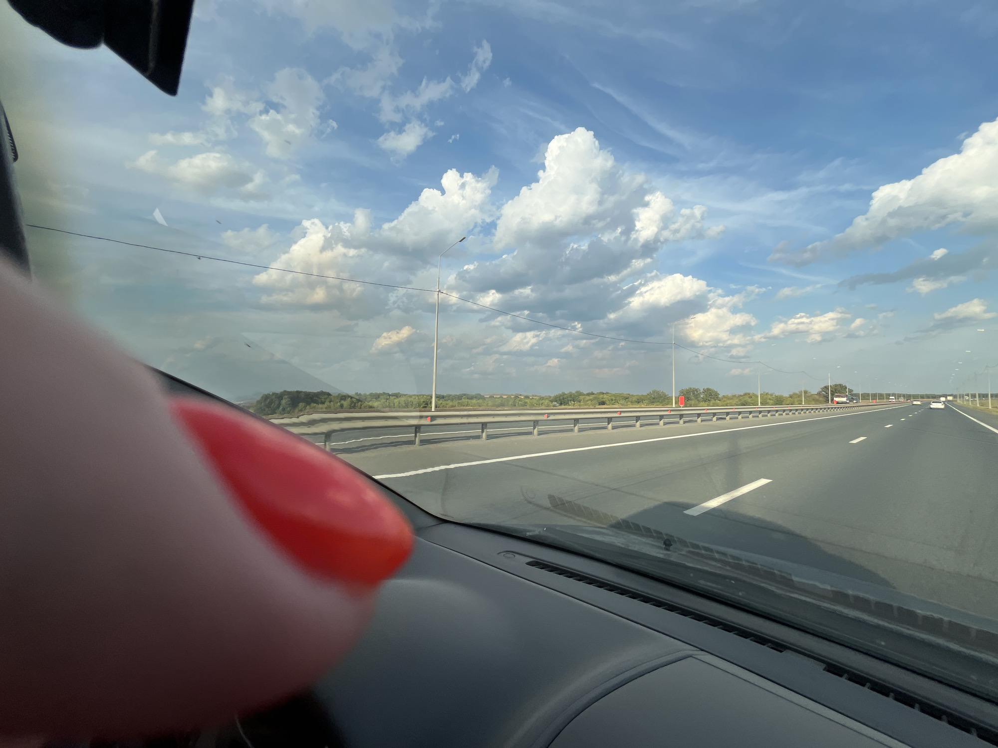 An extreme close-up, POV shot from inside a vehicle shows a bright red fingernail in the foreground, which is out of focus and partially obscuring the view of the windshield. The windshield itself is dirty, with several smudges and debris visible. Beyond the windshield, a highway stretches into the distance under a vast, blue sky filled with dramatic, white cumulus clouds. The highway is empty except for a few distant vehicles. The roadside is lined with trees and vegetation, and there are streetlights visible at intervals. The dashboard of the car is visible at the bottom of the frame. The scene appears to be a snapshot of a road trip or commute, captured from the passenger seat, and there is no clear indication of the specific location in Saratov, Russia, beyond it being a road.