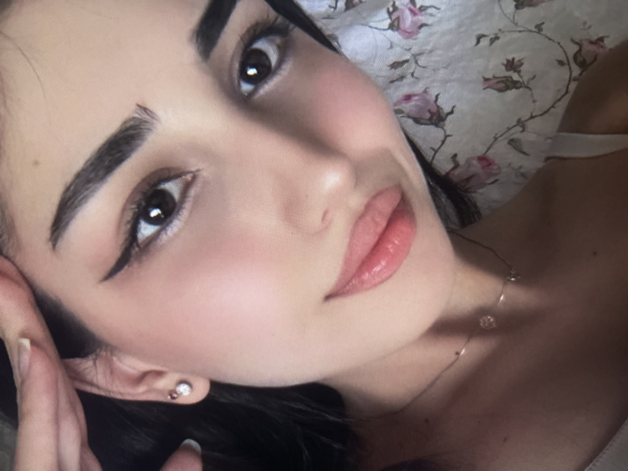 A close-up, slightly blurry, upward-angled selfie shows a young woman with dark hair and dark eyes. She is wearing subtle makeup including eyeliner and lip gloss. Her right ear, adorned with a small stud earring, is visible. A delicate necklace with a small charm rests on her collarbone. She is lying on a floral-patterned fabric, possibly a pillow or blanket. The lighting appears soft and natural. The image does not contain any information about the location being in Qarağandı city administration, Kazakhstan.