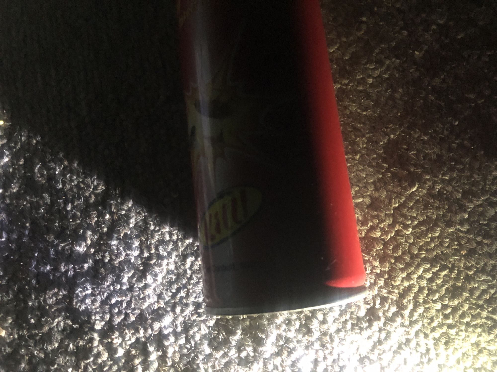 The image shows a close-up, oblique view of a red cylindrical object lying on a textured surface that appears to be a carpet or rug. The object has a label with yellow and red elements and some dark text which is largely obscured by shadow. The lighting in the image is dramatic, with a strong light source creating bright highlights on the surface and casting deep shadows. The surface itself is composed of many small, fibrous elements, with areas of bright illumination and dark shadow, suggesting a plush texture. The image is cropped tightly on the object and its immediate surroundings. There are no people or any indication of the city of Jalingo, Nigeria.