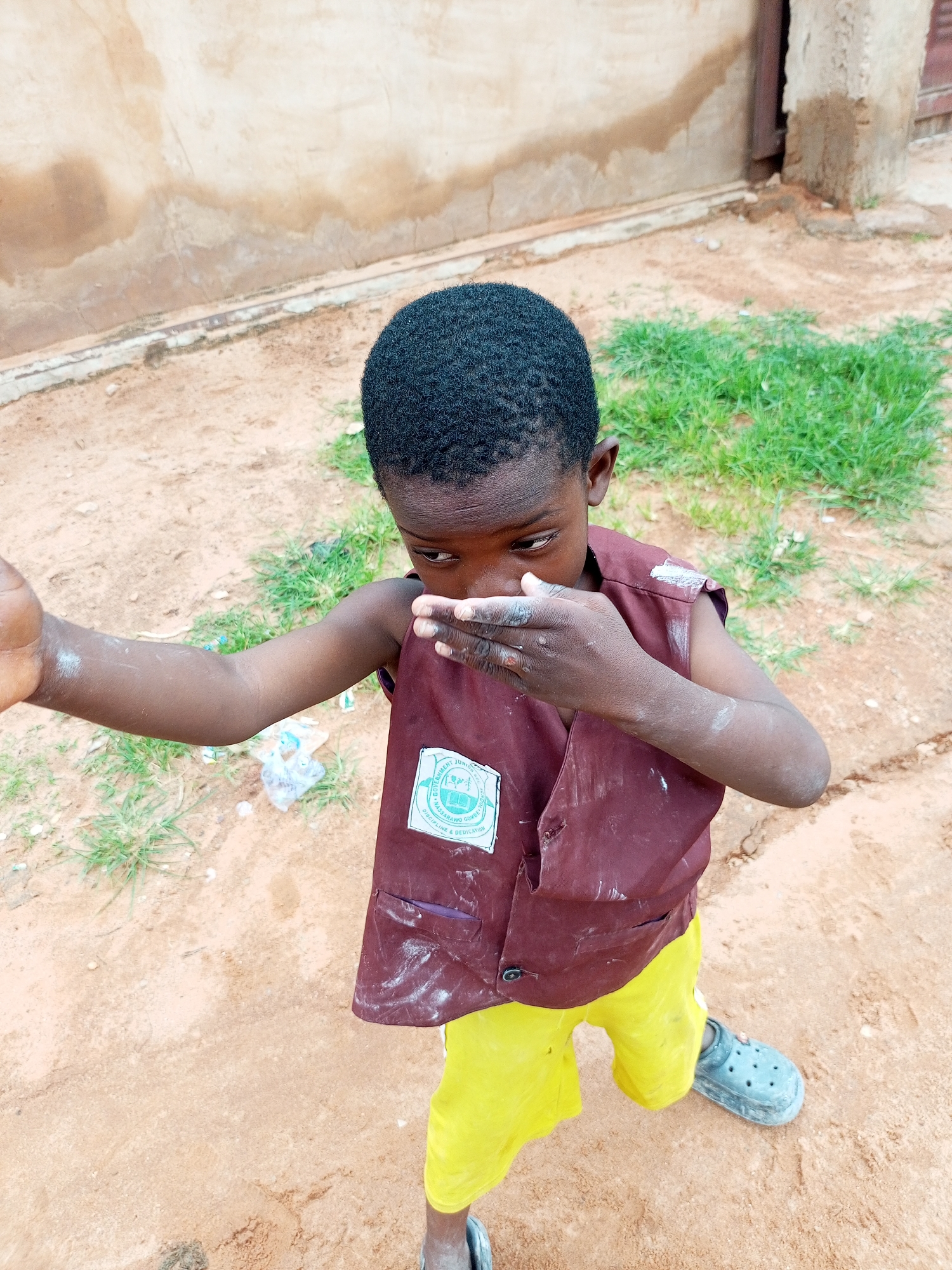 The image shows a young Black boy standing in a sandy outdoor area, likely in Tempure, Nigeria. He is wearing a maroon vest over a yellow shirt and shorts. A white emblem is visible on the left side of the vest, appearing to be a school crest. The boy is holding his hand to his mouth, and there is white powder or dust on his arms and vest. The ground is composed of sand and patches of dry grass. In the background, there is a beige-colored wall, possibly part of a building, with some texture and discoloration. To the right of the wall, a darker vertical structure, possibly a wooden door or frame, is visible. The overall environment suggests a somewhat unkempt outdoor space in a village or urban fringe setting.