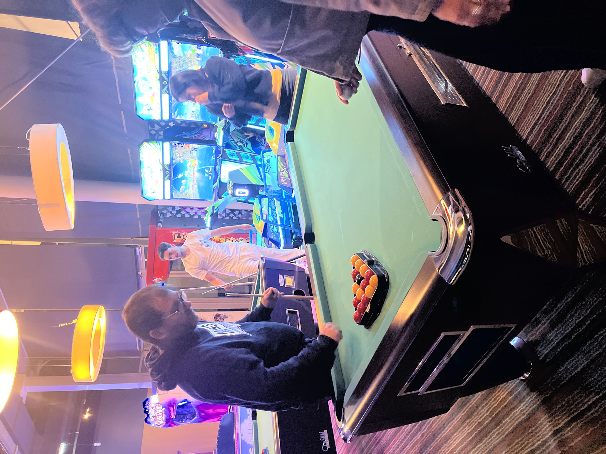 The image depicts an indoor entertainment area, potentially an arcade or entertainment center, in Montrond-les-Bains, France. The scene is dominated by a billiards table, positioned diagonally in the frame, with a full rack of balls. A man wearing a dark hooded sweatshirt is leaning over the table, holding a pool cue. Another person is visible in the background, leaning over an arcade game. Further arcade games are arrayed in the background. Overhead, several yellow ring-shaped light fixtures illuminate the scene. The floor is covered with a brown and tan striped carpet. Other people are standing around, participating in activities near the pool table and in the background.