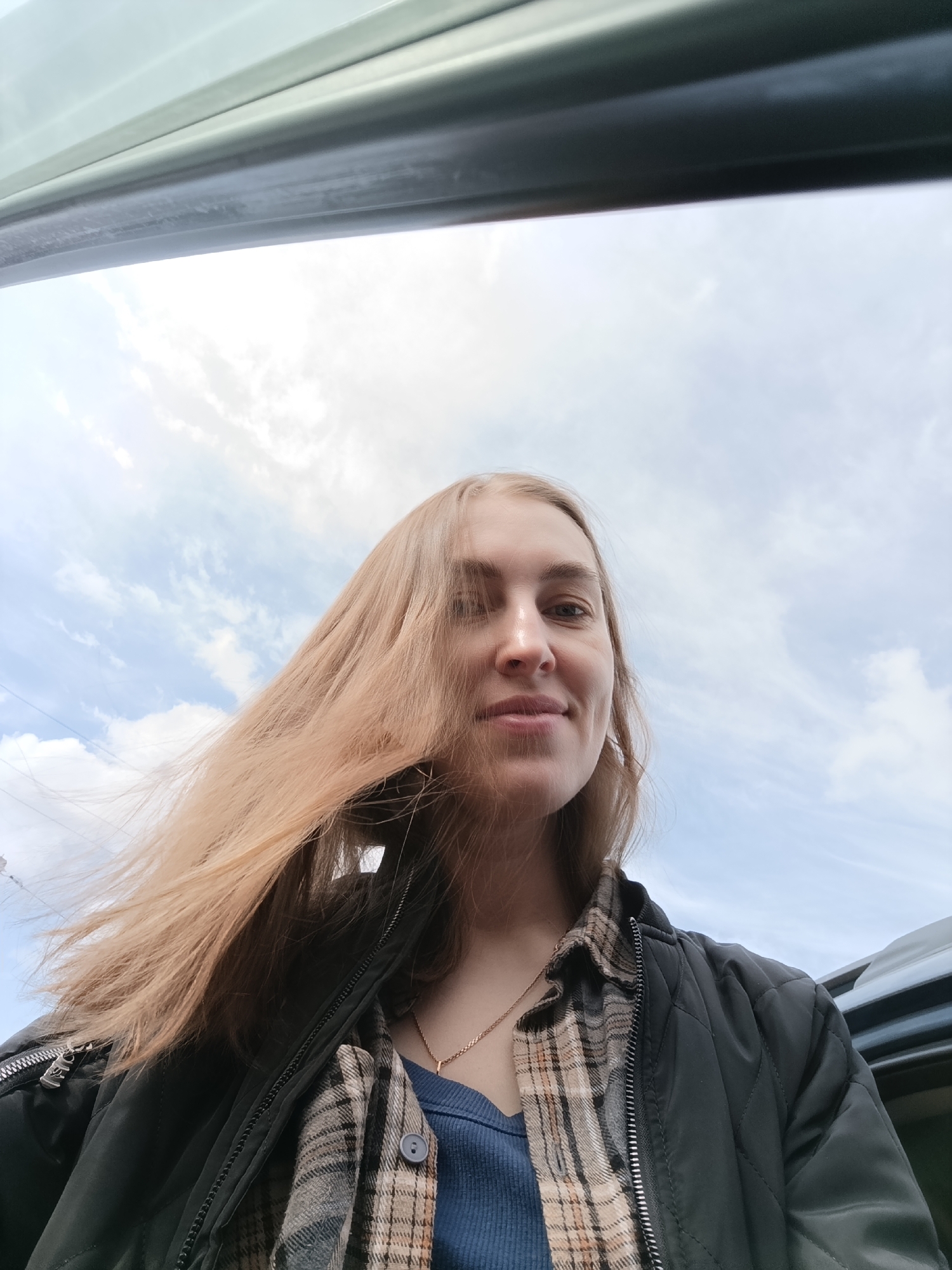A young woman with long, light brown hair is depicted in an upward-angled outdoor photograph. Her hair is windswept across her left side, partially obscuring her face. She has a serene expression with a subtle smile. She is dressed in a black quilted jacket, unzipped, revealing a plaid shirt underneath, which is also open to show a blue ribbed top. A delicate gold-colored chain necklace is visible around her neck.
The background consists primarily of a bright, cloudy sky with numerous white cumulus clouds against a light blue backdrop, indicating daytime. A dark, angled structure, resembling the top frame of a car door or window, spans the upper portion of the image. The scene suggests a casual moment, possibly taken from within or next to a vehicle, amidst a breezy outdoor environment. No discernible text is visible. This scene is set outdoors, potentially in Chaikovsky, Russia.