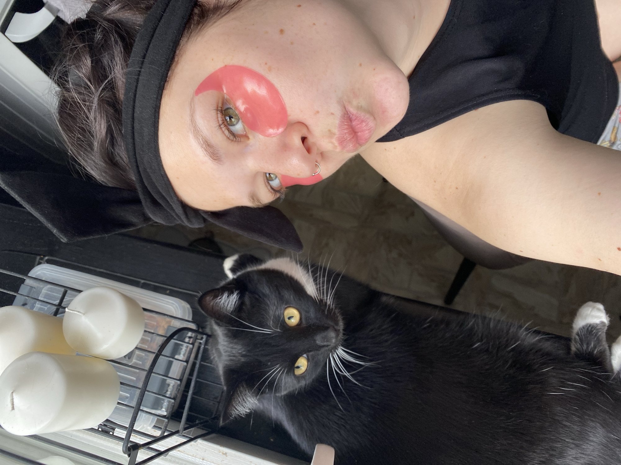 A selfie-style image shows a woman with fair skin and hazel eyes. She has a black headband, and pink under-eye patches are applied to her face. She is wearing a black top. Her nose is pierced with a silver ring. Her lips are slightly pursed.
Beside her is a black and white cat with yellow eyes. The cat is lying down, looking up at the camera.
Behind the woman, there is a black metal shelf holding three white pillar candles and a clear plastic container with a lid. The background is indistinct, but it appears to be an indoor setting.
The location is Chaikovsky, Russia, as indicated by the prompt.