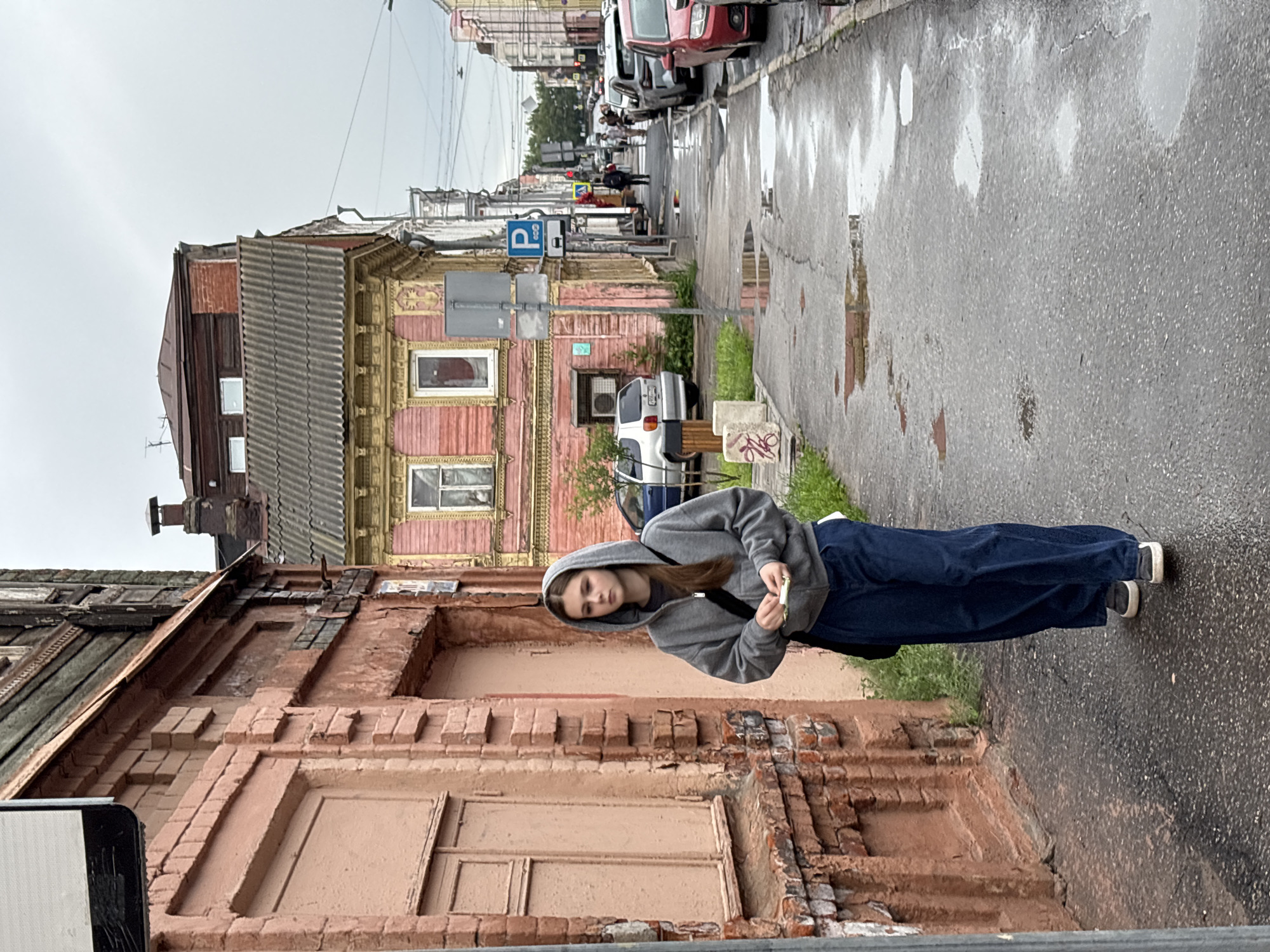 The image, taken in Nizhny Novgorod, Russia, depicts a young person standing on a wet, weathered sidewalk. The person, with long brown hair, is wearing a grey hooded sweatshirt and dark blue pants, with white-soled shoes. They are leaning their head on their hand and holding a small white object. The surroundings suggest a somewhat rundown urban environment. To the left are old, dilapidated buildings with visible brickwork and peeling paint, one appearing to be wooden with ornate trim. To the right, the wet asphalt reflects the overcast sky, with puddles indicating recent rain. In the background, a street is visible with parked cars and a few distant figures. A blue and white parking sign with a "P" symbol is prominent on the left side of the street. The overall impression is of a somber, slightly gloomy day, likely in the afternoon given the light.