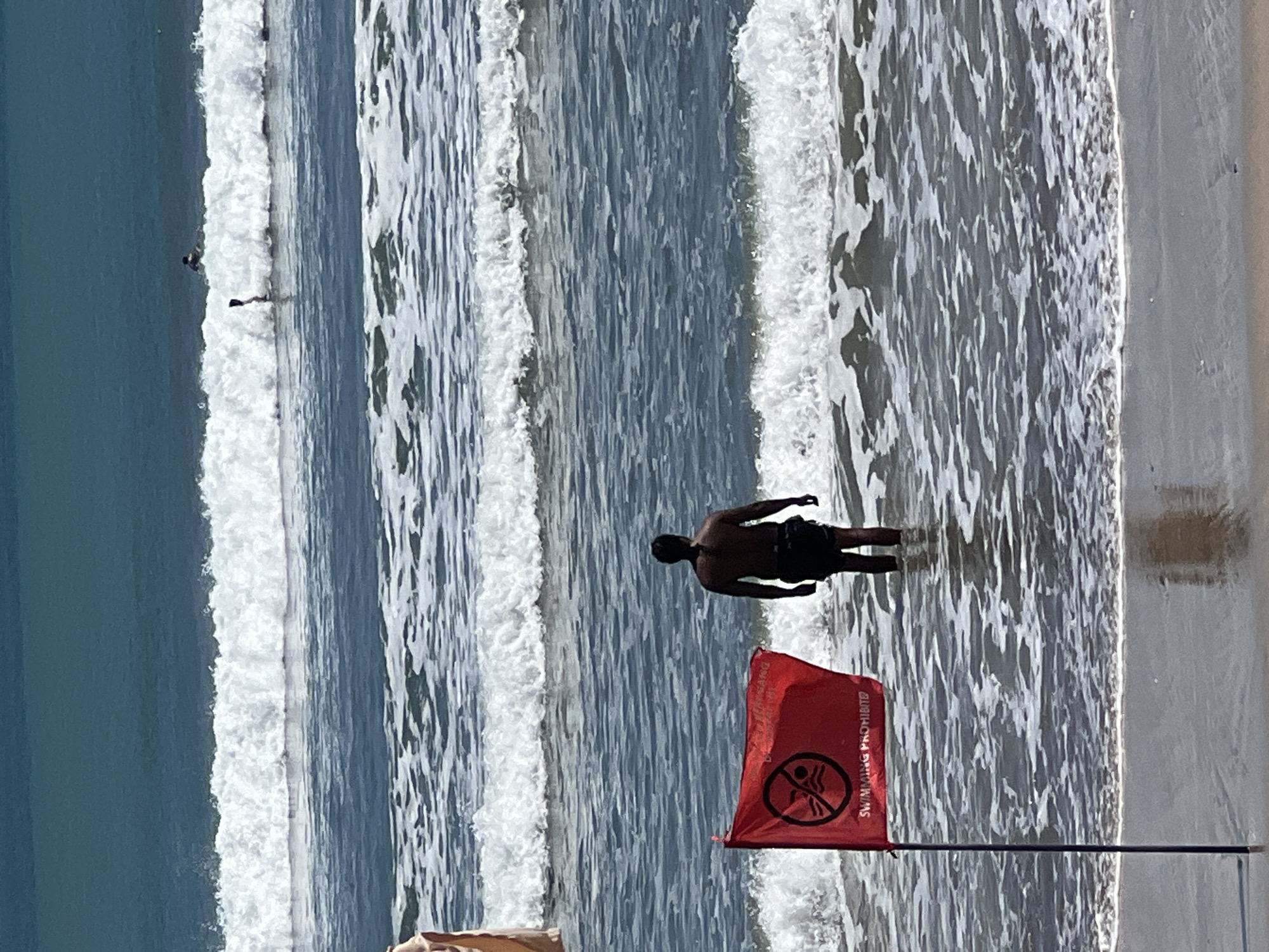 The image shows a man standing in the shallow water of Kuta Beach, Indonesia, facing away from the camera. He is wearing dark swim trunks. In the foreground, a red flag with a white symbol and text is planted on a black pole. The symbol on the flag appears to be a circle with a diagonal line through it, superimposed over a depiction of two people swimming. The text on the flag reads "SWIMMING PROHIBITED." The ocean has multiple rows of white-capped waves. In the distance, two figures are visible on surfboards in the ocean. The sky is a clear, bright blue.