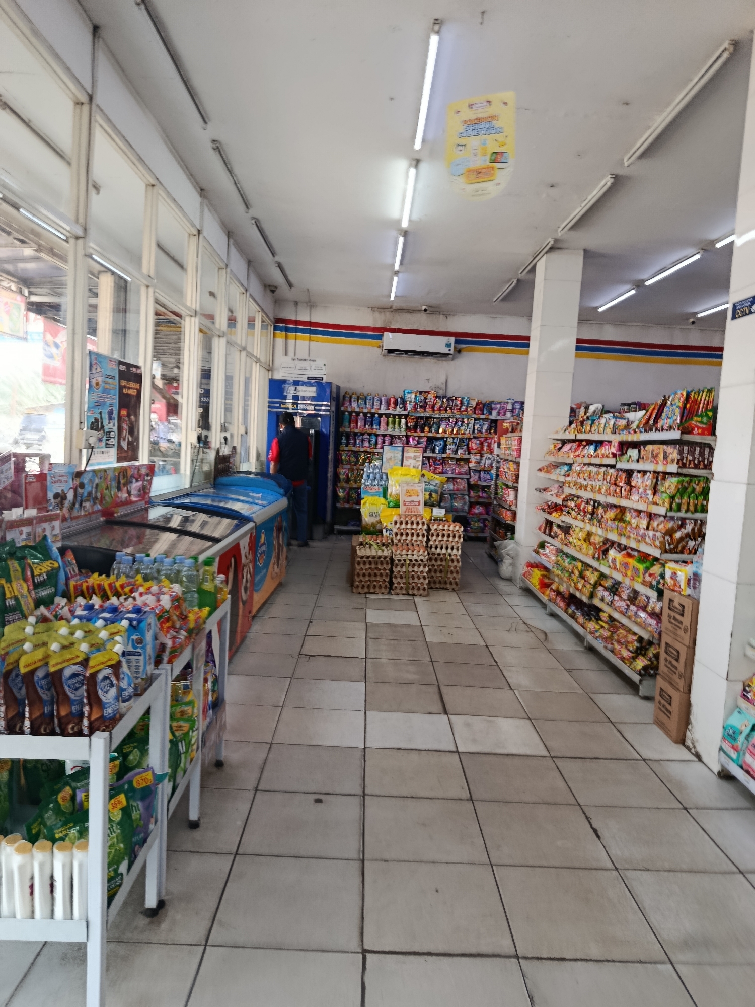 The image depicts the interior of a convenience store, likely located in Wanasari, Indonesia, given the context. The store is well-lit by overhead fluorescent lights. On the left side, a row of refrigerators displays various beverages and frozen goods, with a prominent ice cream freezer. In the center, shelves are stocked with packaged foods and drinks, including many pouches of liquids and boxes of eggs. A person in a red shirt and dark pants is visible standing near a blue vending machine or payment station. Towards the right, tall shelves are filled with a wide assortment of snacks and other packaged consumer goods. The floor is tiled with light-colored square tiles. A yellow promotional banner is hanging from the ceiling in the upper portion of the image. The store appears to be open for business, with merchandise neatly arranged.