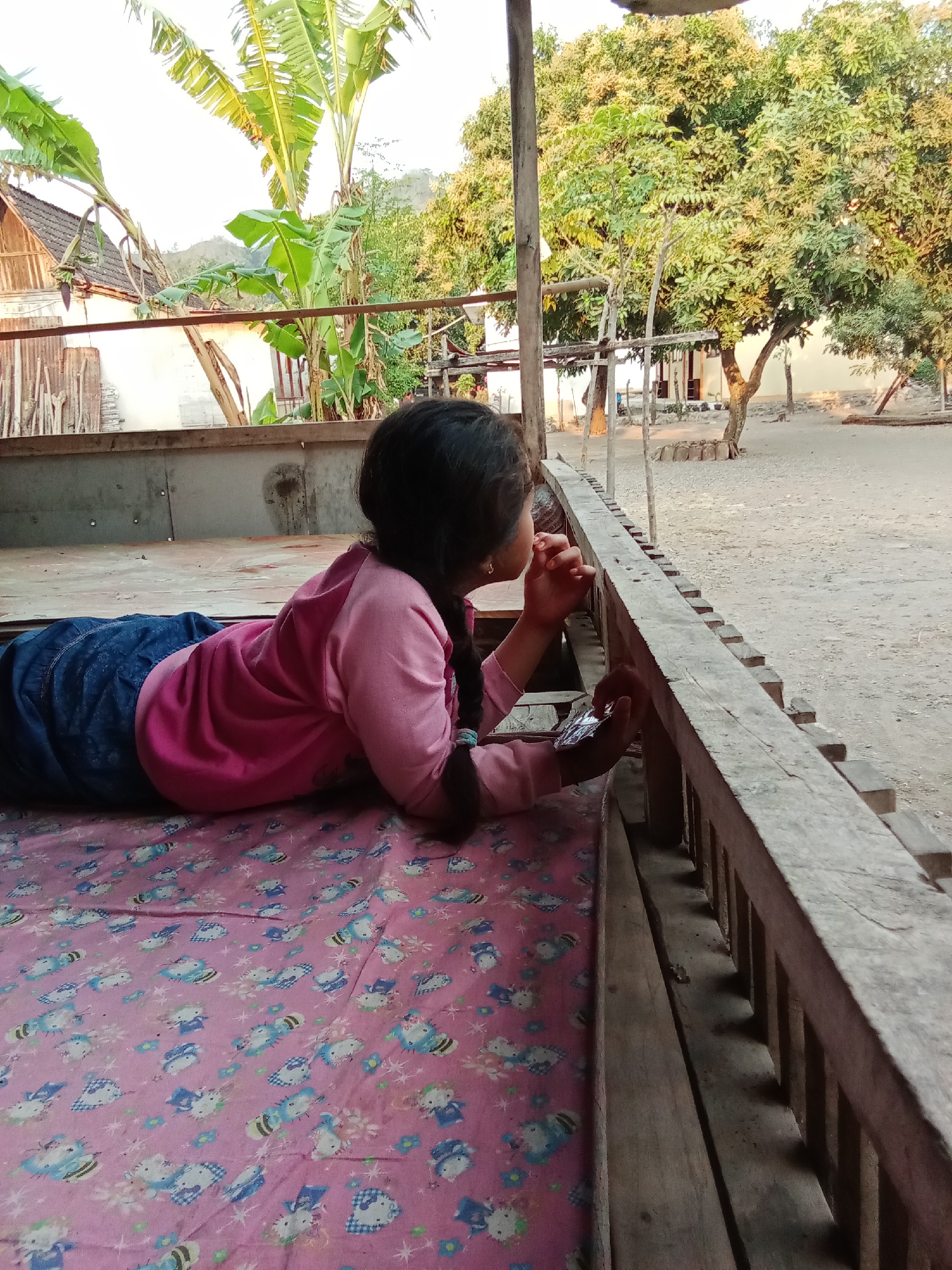 A young girl with dark hair braided down her back is lying on her stomach on a pink mattress adorned with cartoon cats. She is wearing a pink long-sleeved shirt and blue pants. Her elbows are on the mattress, and her hands are clasped in front of her face. She is looking out towards a wooden railing and beyond, where a dirt ground and trees are visible. In the background, there are banana trees, a building with a tiled roof, and more trees. The lighting suggests it is daytime, possibly late afternoon. The scene appears to be outdoors, in a rural or semi-rural setting. This image was taken in Selowogo, Indonesia.