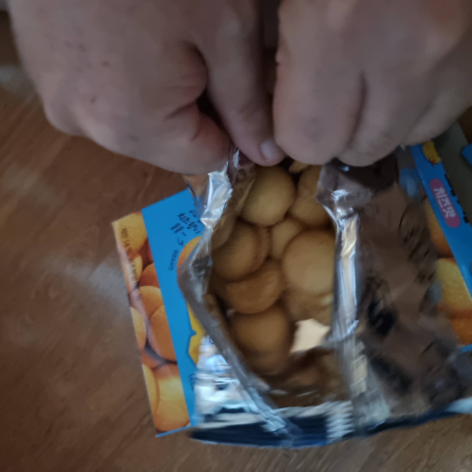 A close-up, eye-level shot shows a person's hands opening a package of small, round, golden-brown baked goods, possibly cookies or crackers. The package is partially open, revealing the contents inside. The baked goods are densely packed within a silver foil bag. The outer packaging, visible in the background and at the bottom of the frame, is a blue cardboard box with some text and images of the baked goods. The scene appears to be indoors, with a wooden floor visible in the background. The lighting is somewhat dim, creating shadows.