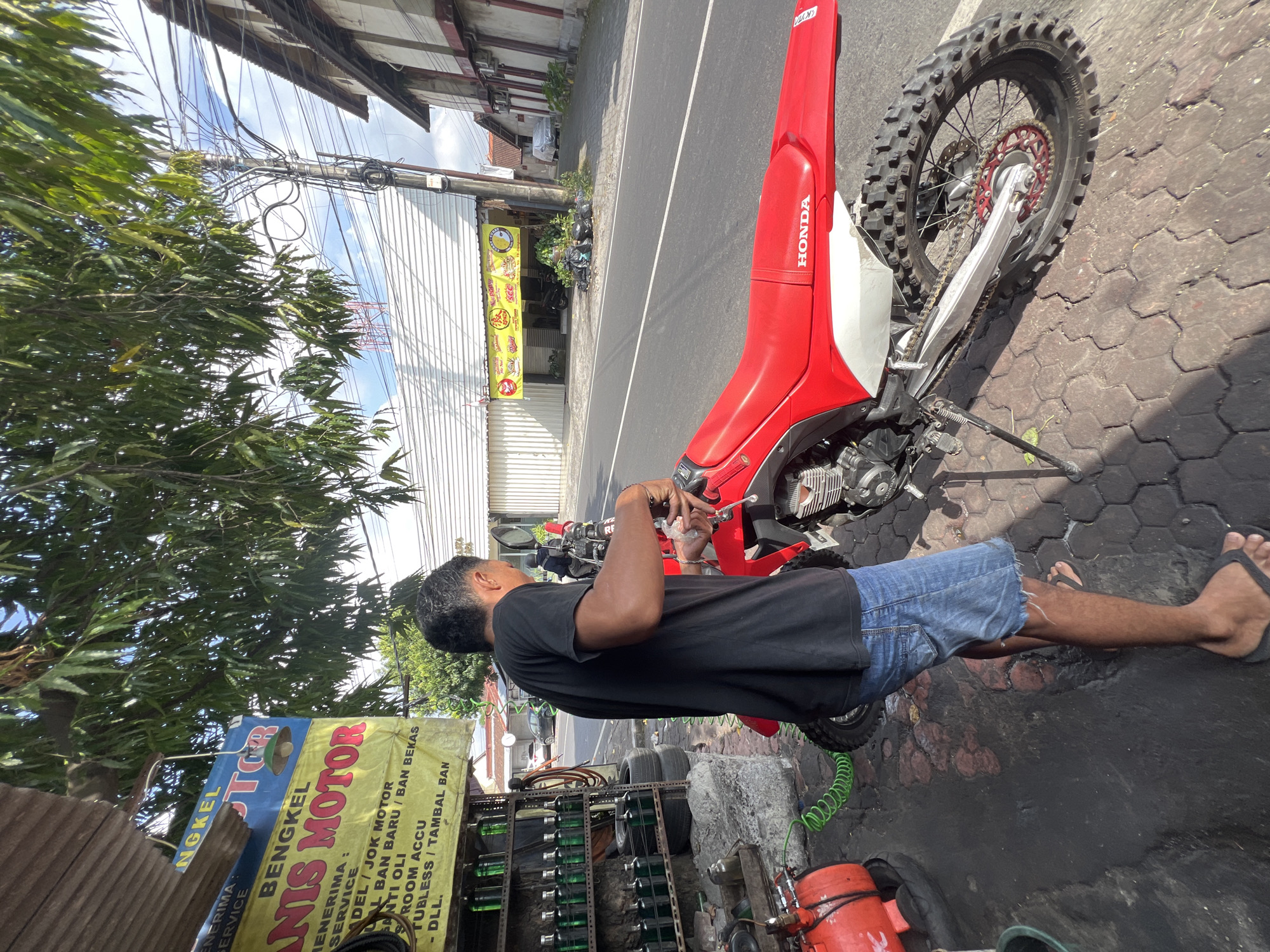 A man in a black t-shirt and denim shorts is standing next to a red Honda dirt bike. He appears to be working on the motorcycle's handlebars. The scene is in front of a small shop with a sign that reads "BENGKEL MOTOR". The shop has shelves stocked with bottles of oil and other supplies. There is also an air compressor and various tools visible. The street in front of the shop is paved with hexagonal tiles. In the background, there are buildings and power lines, suggesting an urban environment. The overall impression is that of a motorcycle repair shop on a street in Denpasar, Indonesia.