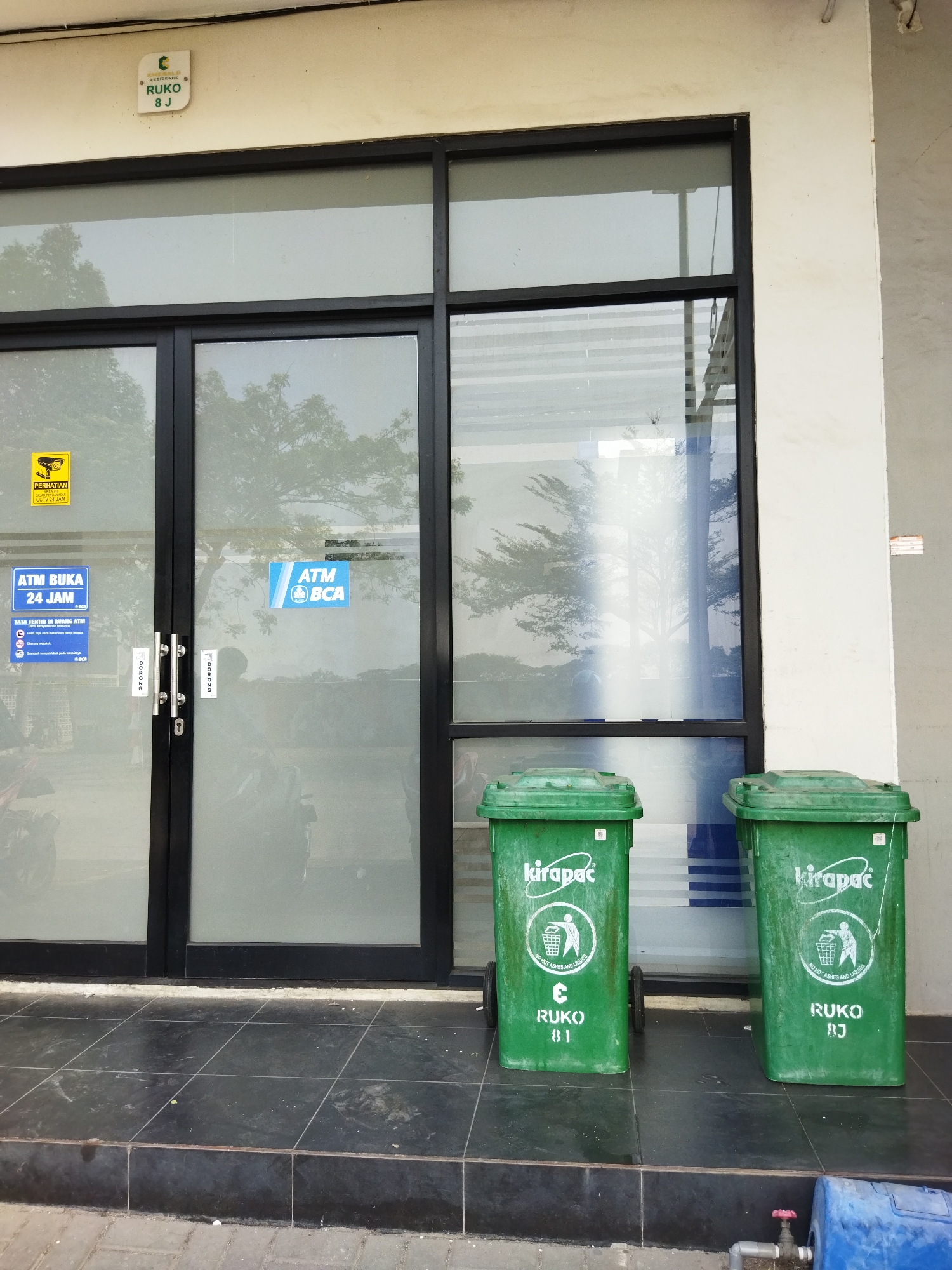 In Kosambi, Indonesia, a commercial building features a storefront with large glass doors and windows. A sign above the entrance indicates "RESIDENCE RUKO 8 J". On the left door, a yellow sign reads "PERHATIAN CCTV" with a camera icon, indicating surveillance. Below this, a blue sign states "ATM BUKA 24 JAM" and "TATA TERTIB DI RUANG ATM". On the right door, a blue sign displays "ATM BCA". Two green trash bins, labeled "kirapac" and "RUKO 81" and "RUKO 85", are positioned outside the entrance. The ground is tiled in black and grey.