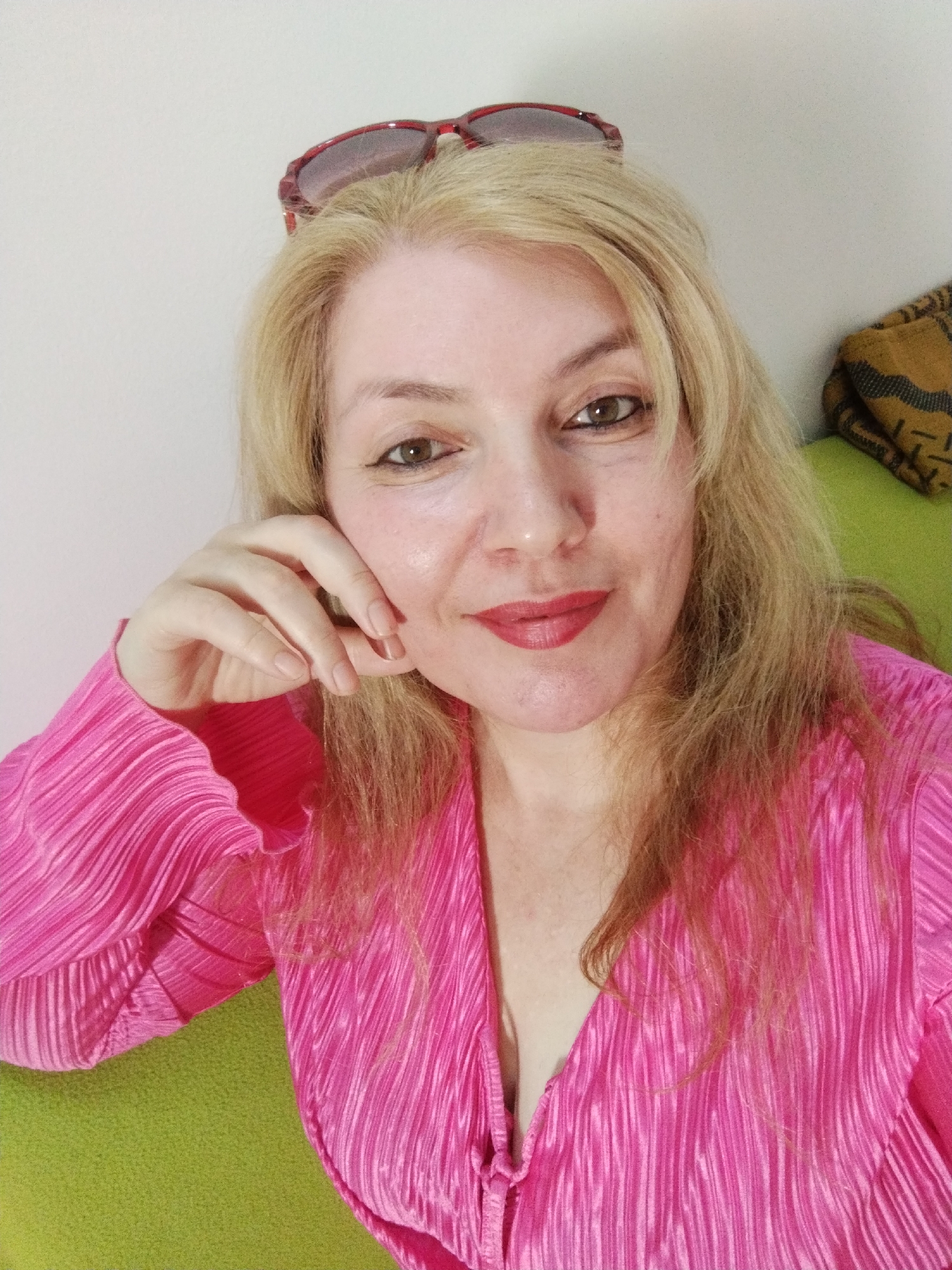 A fair-skinned woman with long blonde hair and dark brown eyes is posing for a selfie. She is wearing a bright pink pleated top with a low V-neck and has red sunglasses perched on her head. Her left hand is resting against her cheek. She is sitting on a green upholstered surface in front of a white wall with a brown and yellow patterned textile visible in the top right corner. The location is not discernible from the provided image.