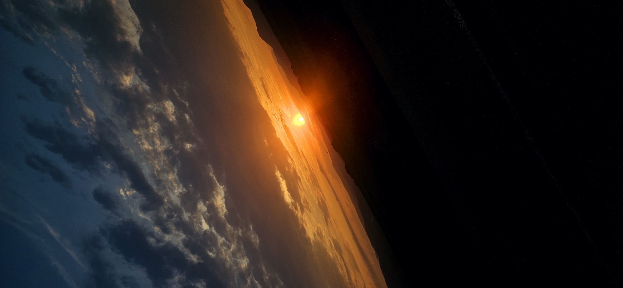 The image displays a diagonal sunset scene over a natural landscape, rotated such that the horizon line runs from the top-right to the bottom-left. The location is Momin Prohod, Bulgaria. The upper-left quadrant of the sky is a dark blue, transitioning through shades of grey and purple to brilliant orange and red hues towards the sun. Clouds are present across the sky, appearing as dark grey formations with edges illuminated in golden and orange tones, particularly near the light source. The sun, a prominent, intense yellow-orange orb, is positioned approximately one-third down from the top edge and slightly to the right of the vertical center, casting a strong radiant glow. Below the sun, a layered series of dark grey to black mountain or hill ranges forms the horizon, silhouetted against the vibrant sky. The bottom half of the image is an undifferentiated black mass, representing the immediate foreground, with no discernible details due to extreme underexposure. No human figures, animals, or man-made objects are identifiable within the frame.