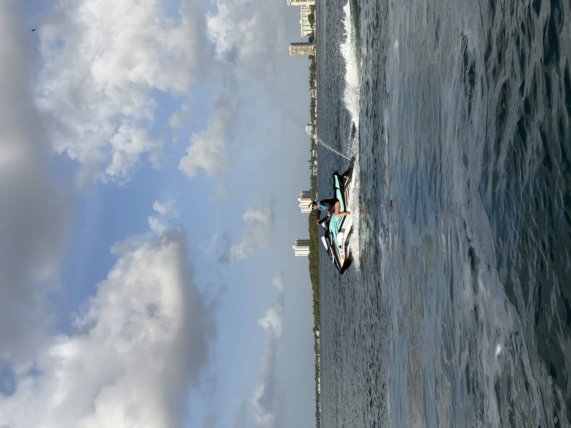 The image depicts a jet ski operating in the waters off the coast of North Miami, Florida. A person is riding the jet ski, which is creating a spray of water as it moves. The shoreline of North Miami is visible in the background with buildings. The sky is partly cloudy with patches of blue. The water appears choppy.