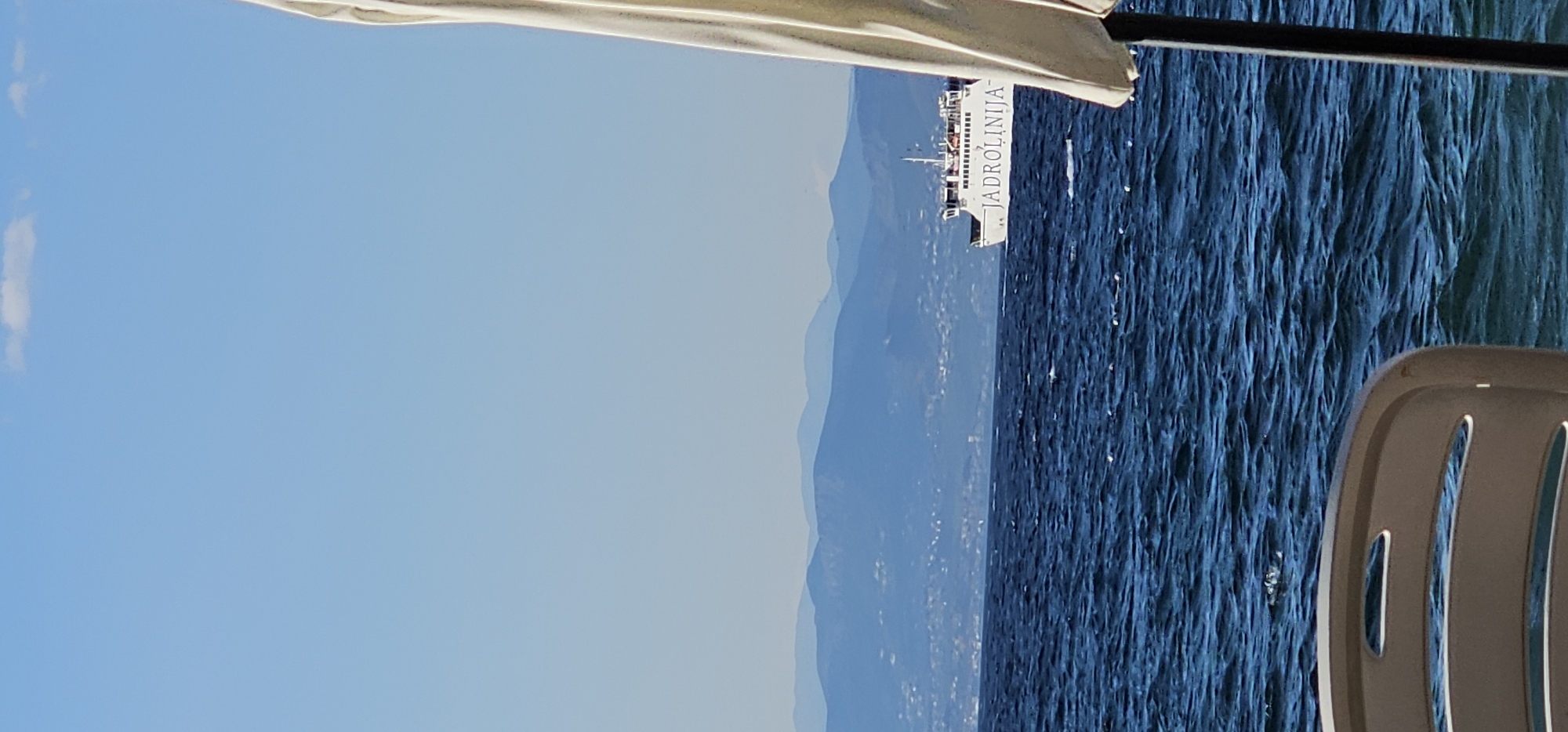 A white ferry with the name "JADROLINIJA" printed in black letters on its side sails on a deep blue sea under a clear sky. The ferry is in the middle ground of the image, moving from right to left. In the background, a range of hazy blue mountains stretches across the horizon, with a small settlement visible on the slopes. The foreground is dominated by the choppy surface of the sea, and a portion of a white beach umbrella and a light-colored plastic chair are visible on the right and left sides of the image respectively. The scene appears to be a sunny day at a coastal location in Supetar, Croatia.
