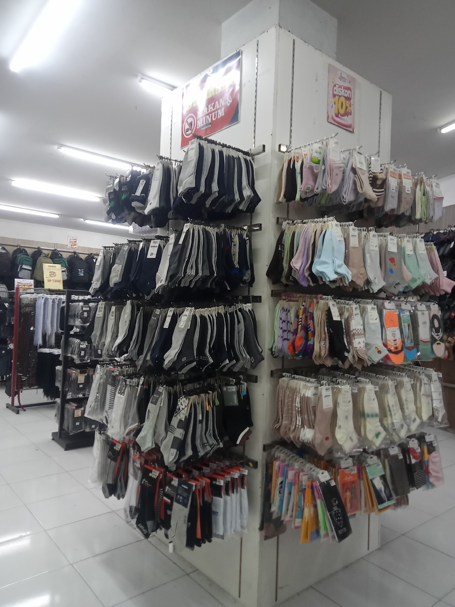 The image depicts a retail display of socks and other apparel in a store located in Boyolali, Indonesia. The display is organized on vertical shelves and hanging racks, showcasing a wide variety of socks in different colors, patterns, and packaging. The socks are predominantly displayed in pairs, with many featuring individual plastic packaging or are hung by clips.
Promotional signage is visible, including a red sign with white text that reads "DILARANG MAKAN & MINUM" (No Eating & Drinking) and another red sign with white text indicating "Diskon 10%" (10% Discount). There is also a yellow sign with "Rp 7.500" suggesting a price point.
In the background, various bags, possibly backpacks, are displayed on a shelf. The environment appears to be a well-lit retail space with white tiled flooring and fluorescent lighting. No individuals are clearly visible in the frame. The overall impression is that of a commercial setting within a store in Indonesia.