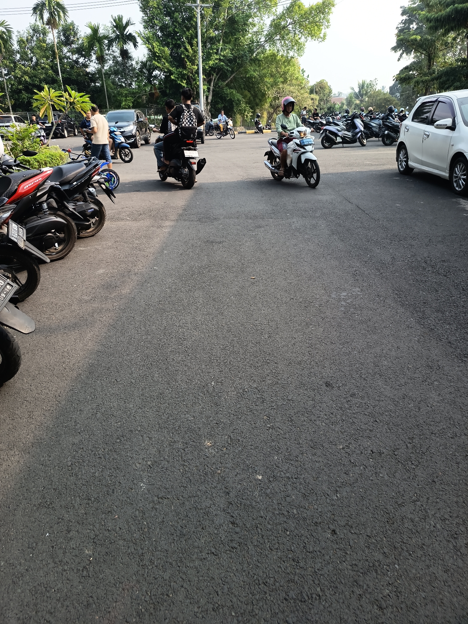 The image depicts a parking area with numerous motorcycles and a few cars. The ground is paved with asphalt. In the foreground, several parked motorcycles are visible. A white car is parked on the right side of the frame. In the mid-ground, a person is riding a white scooter with a pink helmet. Further back, more parked motorcycles are visible, and some people are standing near them. The background features lush green trees and vegetation, suggesting an outdoor location. The overall scene appears to be a typical parking lot in a city, possibly a public area or a commercial establishment. The specific location in Lubuklinggau, Indonesia, cannot be definitively determined from the visual information alone, as there are no recognizable landmarks or signs specific to that city. However, the presence of many motorcycles and the tropical foliage are consistent with an Indonesian environment.