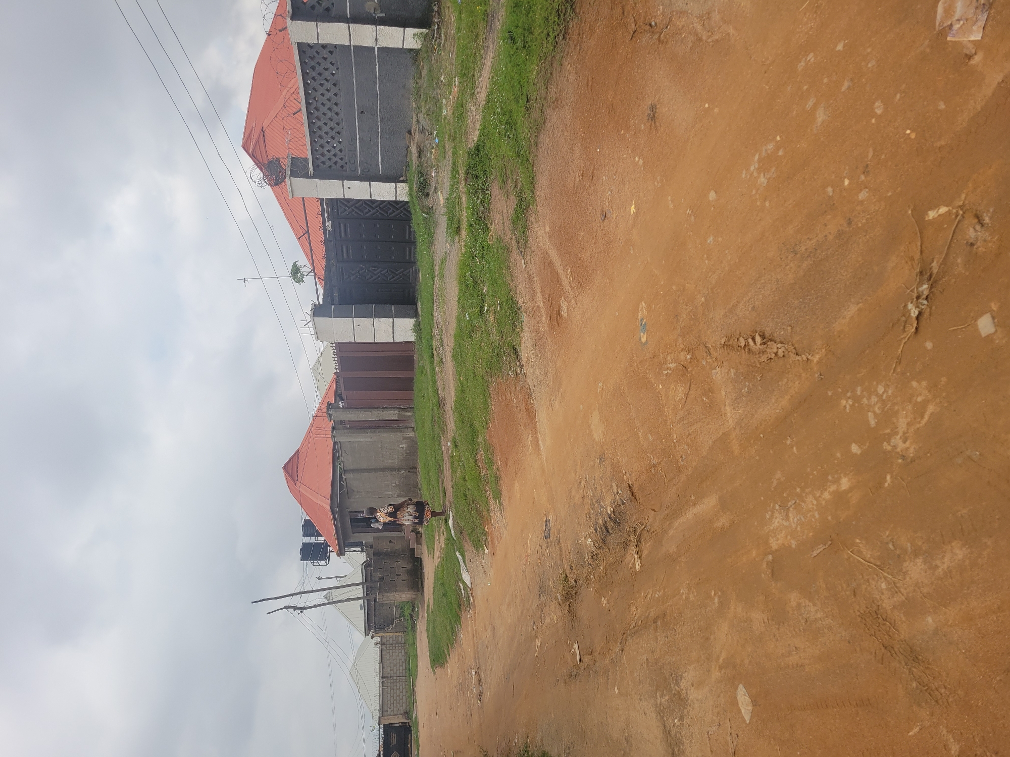 This is a medium shot, eye-level, exterior photograph taken on a cloudy day in Madalla, Nigeria. The foreground is dominated by a dirt road, which appears worn and has some tire tracks and debris. In the middle ground, on the right side of the road, is a building with a red, conical roof and concrete walls. A person in a patterned dress stands in front of this building. To the right of this building is a dark metal gate with decorative patterns, flanked by white and grey pillars. Beyond this, on the far right, is a section of a grey wall with a lattice pattern. On the left side of the road, in the middle ground, are other buildings with corrugated metal roofs and concrete walls, some with barbed wire on top. Utility poles and power lines are visible across the upper portion of the image. The overall environment suggests a residential area with unpaved roads.