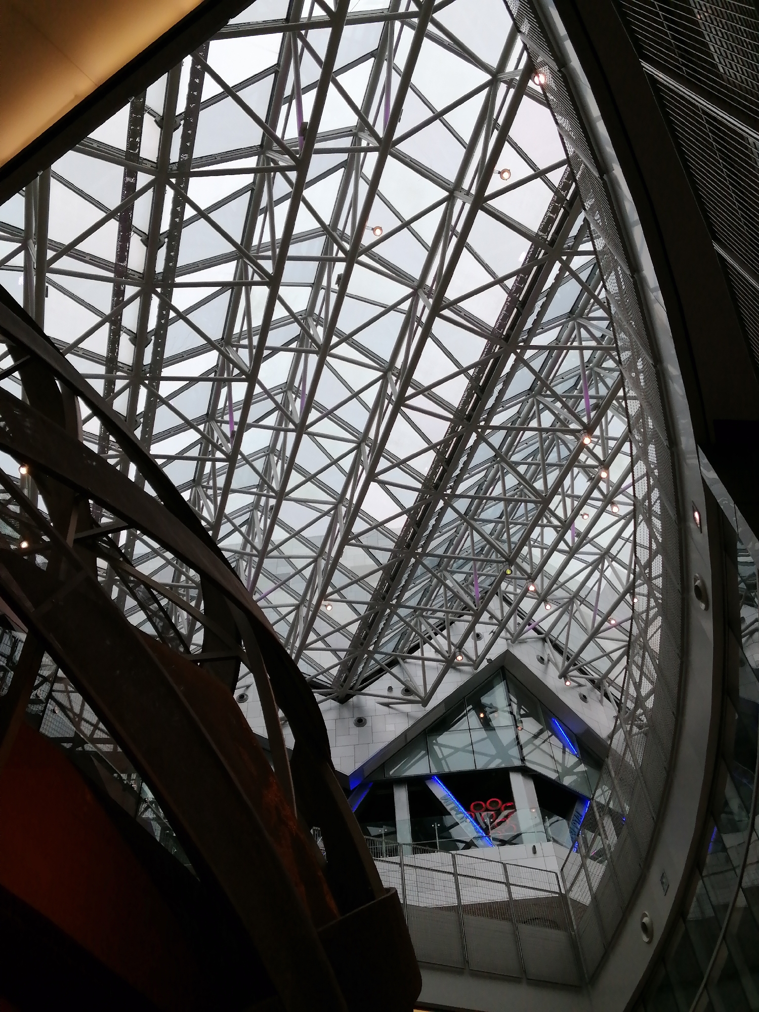 The image shows an upward view of a complex architectural structure. A web of intersecting metal beams and glass panels forms a geometric pattern across the ceiling, suggesting a modern building. In the foreground, dark, curved metal beams create an abstract, artistic element within the frame. A multi-level building with glass windows and blue lighting is visible, with what appears to be a red neon sign. The scene takes place inside a building. The image possibly depicts the interior of the Afimall City shopping center in Moscow, Russia.