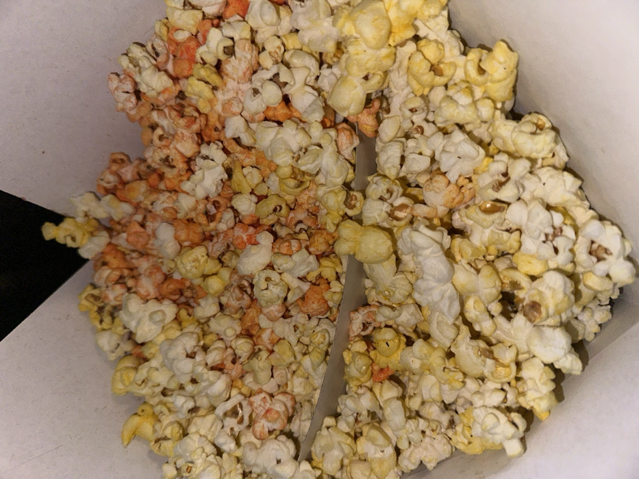 The image displays a close-up, top-down view of popcorn within a white, two-compartment cardboard container. The popcorn consists of various kernels, predominantly white and yellowish, intermixed with numerous pieces coated in a reddish-orange seasoning, indicative of chili powder. A cardboard divider bisects the container, separating the popcorn into two distinct portions. The background is dark, suggesting an indoor environment, likely a cinema in Mexico City, Mexico. No individuals or specific actions are visible.