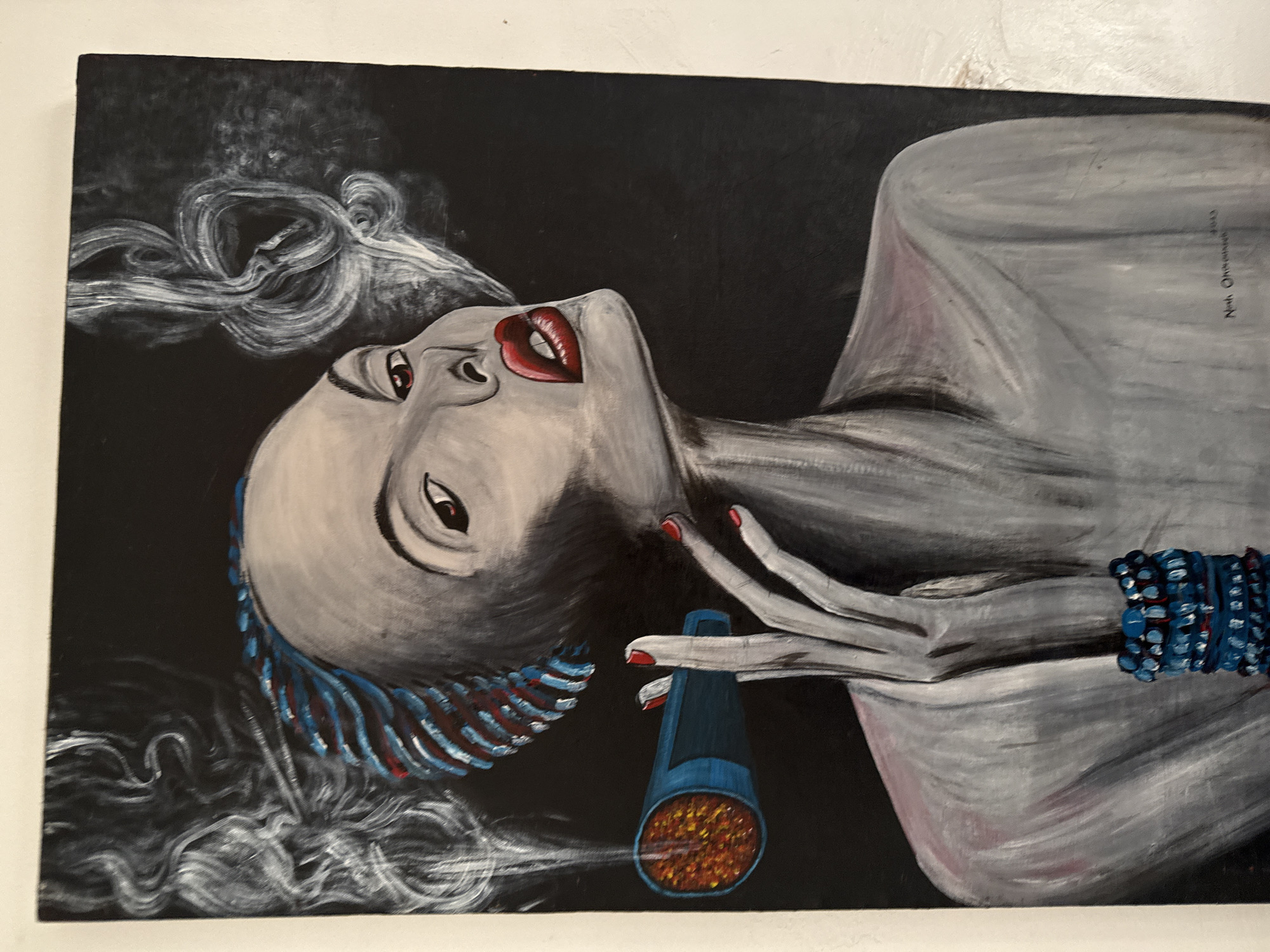 The media file displays a stylized painting of a woman, rendered primarily in shades of grey and white against a stark black background. The woman is depicted from the chest up, reclining or looking upwards with an intense gaze. Her lips are a striking vivid red, and her eyes feature subtle red accents. Her hair, which appears textured or braided, incorporates streaks of blue and red, and merges into swirling white smoke that billows around her head.
The woman holds a dark blue pipe-like object with red detailing in her right hand, from which glowing orange and yellow embers are visible. Her left wrist is adorned with a thick, multi-strand bracelet made of small blue beads.
The overall scene captures a moment of contemplation or reverie, with the smoke adding to the dramatic and expressive quality of the artwork. The painting utilizes strong contrasts between the monochromatic figure and the vibrant accents of red, blue, and the glowing embers. The artistic style combines recognizable features with expressive, almost flowing lines, particularly in the smoke and hair.
Visible text on the upper right side of the painting reads "Nne Onyeamara 2023", indicating the artist's name and the year the artwork was created.