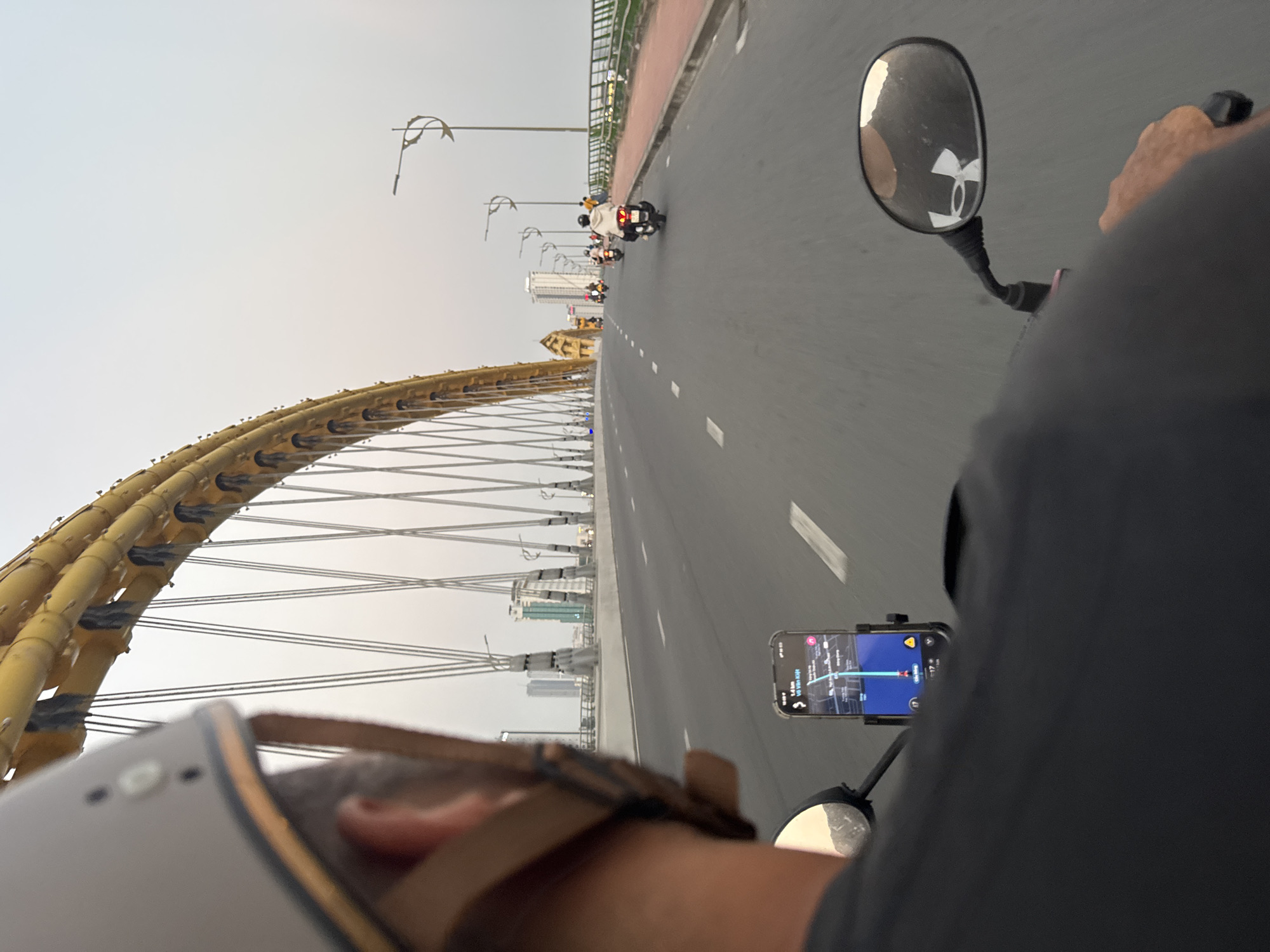 The image depicts a first-person perspective from a motorcycle riding across the Dragon Bridge in Đà Nẵng, Vietnam. The yellow dragon structure of the bridge is visible on the left. The road is divided by white lines, with other motorcycles ahead. The rider has a phone mounted, displaying a navigation app, and is wearing a helmet. A side mirror is visible, reflecting the Under Armour logo. The sky is overcast.