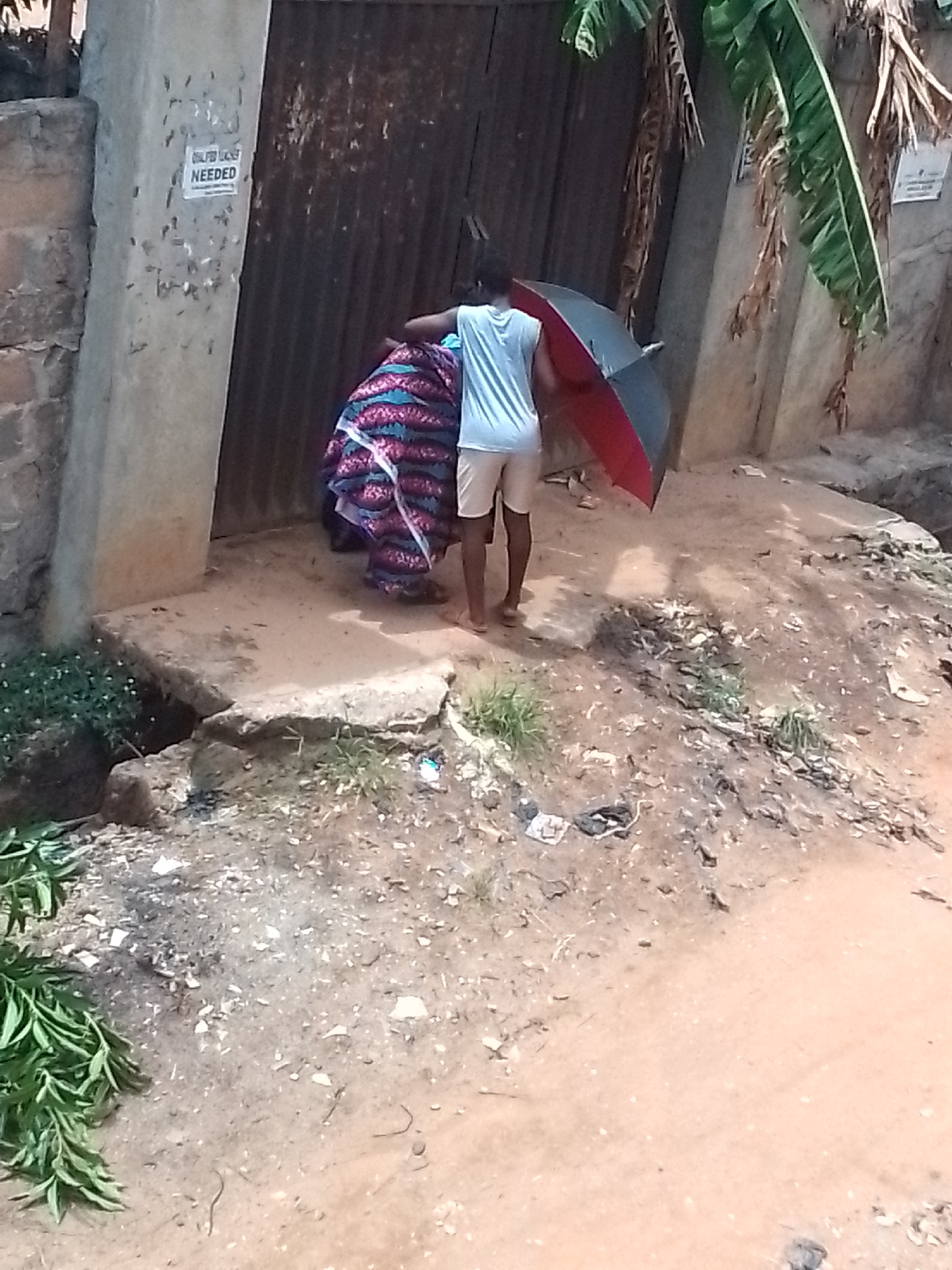A man holding an open, black and red umbrella is helping a woman with a large, multicolored patterned burden through an open gate, with a sign that reads "SAFE LICE NEEDED" affixed to the wall adjacent to the gate. The scene includes sparse vegetation and trash-scattered, bare earth.
