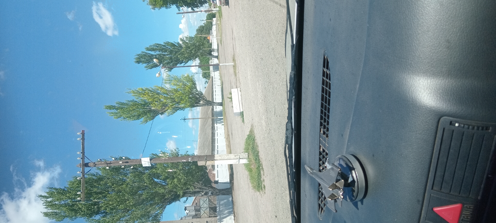The image shows a view from inside a vehicle looking out at a street scene in Koviahy, Ukraine. The sky is bright blue with scattered white clouds. Tall, slender trees line the street, with one prominent utility pole in the foreground supporting power lines. In the background, a long, low building with a grey roof is visible, bordered by a white fence. A small concrete planter box sits on the side of the road. The interior of the vehicle is visible in the foreground, with a dashboard featuring a vent and a small model airplane on a stand. The model airplane is a silver jet with its wings spread. The overall scene depicts a typical day in a semi-rural or industrial area.