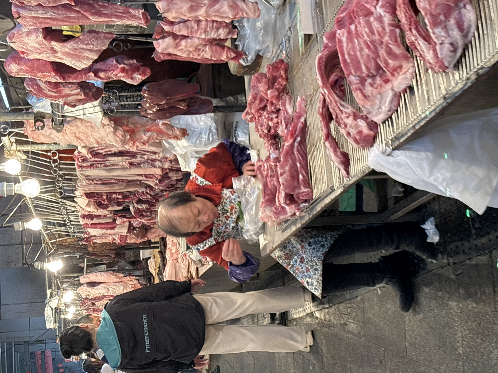 The image depicts a butcher shop in Jiangjin, China. An elderly woman in a red jacket and floral apron stands behind a counter laden with various cuts of raw meat, likely pork, hanging above and displayed on the surface. She appears to be interacting with the meat. A person wearing a black jacket with the word "PNDEPENDENCH" on it and light-colored pants stands facing the butcher, presumably a customer. Overhead, rows of hanging meat are illuminated by bare light bulbs. The surrounding environment appears to be an open-air market, with a concrete floor visible.