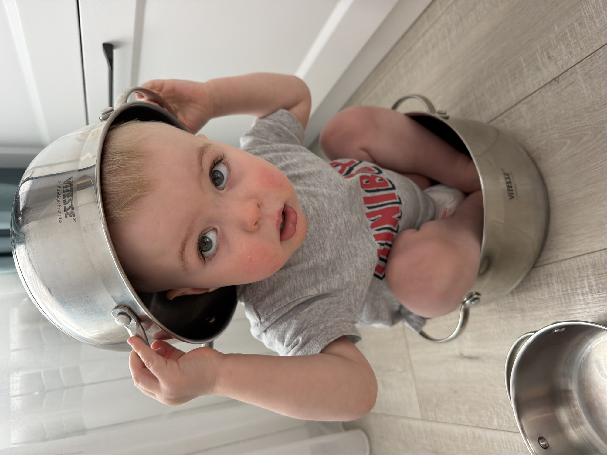 A baby is sitting in a silver pot on a wooden floor. The baby is wearing a grey shirt with red lettering and has another silver pot on its head. The baby has blonde hair and blue eyes and is looking at the camera with its mouth slightly open. In the background, there is a white cabinet. There is no indication of the location in Votkinsk, Russia.