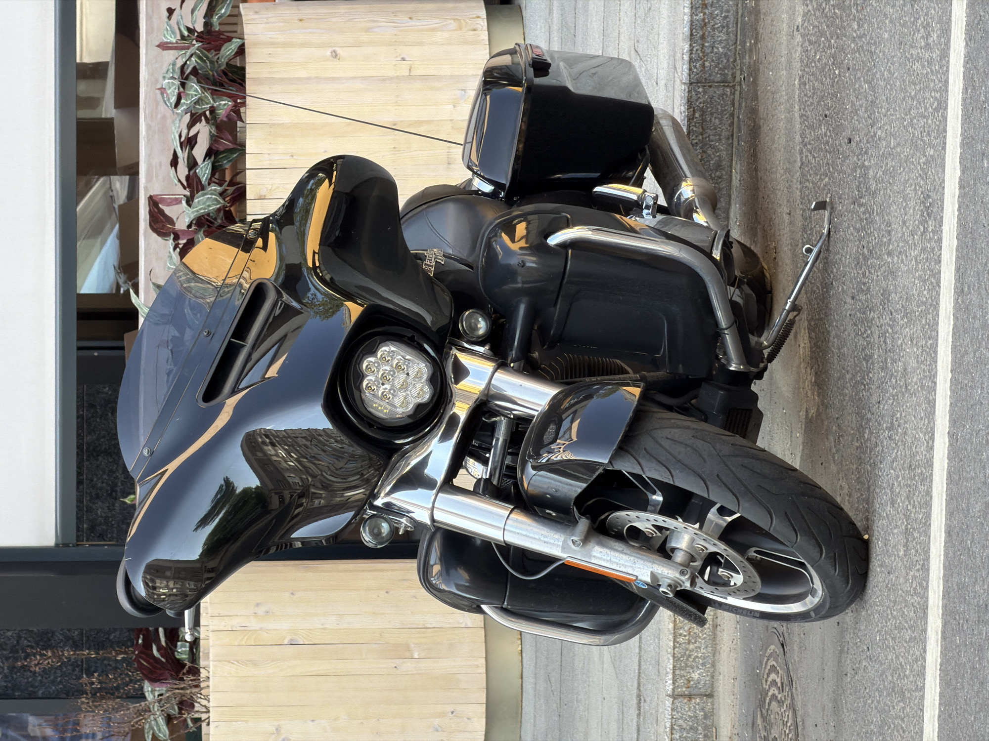 A black motorcycle is parked on a sidewalk in Moscow, Russia. The motorcycle is positioned near a building with a wooden facade and greenery. The front tire faces the street with a white line marking the road. The reflection on the motorcycle's fairing reveals the surrounding urban environment.