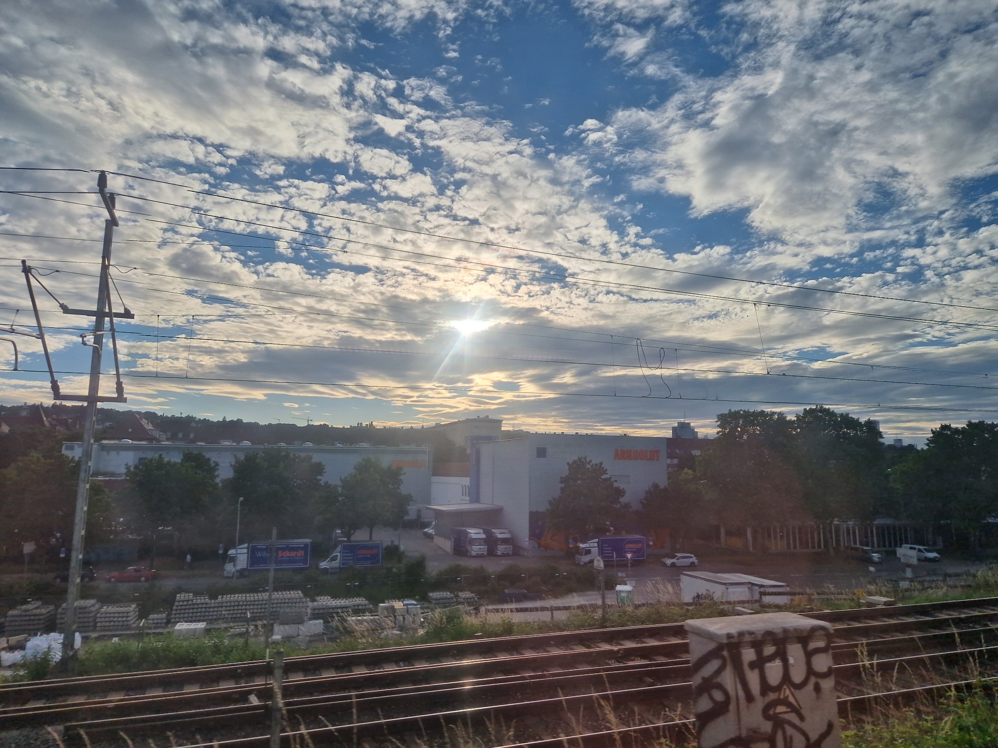 The image depicts a view from a train track in Stuttgart, Germany, looking towards a commercial area under a cloudy sky with the sun shining through. In the foreground, there are multiple train tracks with grass and weeds growing between them. A concrete utility box with graffiti is visible in the lower right corner.
In the middle ground, there is a road with several vehicles, including trucks and cars. One truck has the text "Wilh. Scharidt Stuttgart" visible on its side. There are stacks of construction materials, possibly concrete blocks, and white bags near the road.
Further in the background, there is a large commercial building with the text "ARNOLDT" and "Stuttgart" on its facade, indicating it is likely a store or distribution center. Trees line the area in front of the building. The sky is filled with dynamic cloud formations, with the sun creating a bright glare. In the far distance, buildings and urban structures of Stuttgart are visible, blending into the horizon. The overall impression is a view of an industrial or commercial zone on the outskirts of the city, seen from the perspective of a passing train.