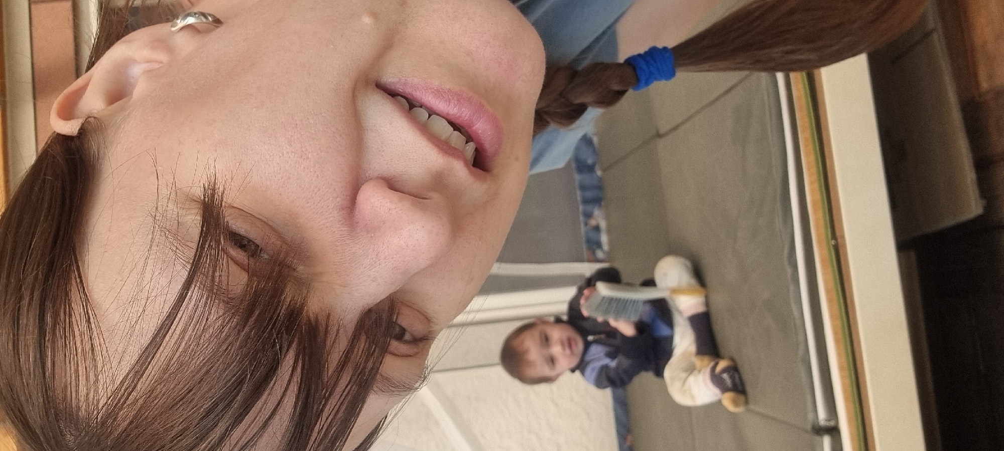 A selfie-style image presents an adult female face at a sharp angle. Her complexion is fair, and her brown hair hangs partially across her face. She appears to have a braid tied with a blue band. Reflected in the backdrop is a young child, likely a toddler, sitting on the floor and playing with a brush. The background also includes some undefined objects and a glimpse of what seems to be the floor. Based on the Korean characters visible, the location is Dunnae-myeon, South Korea.