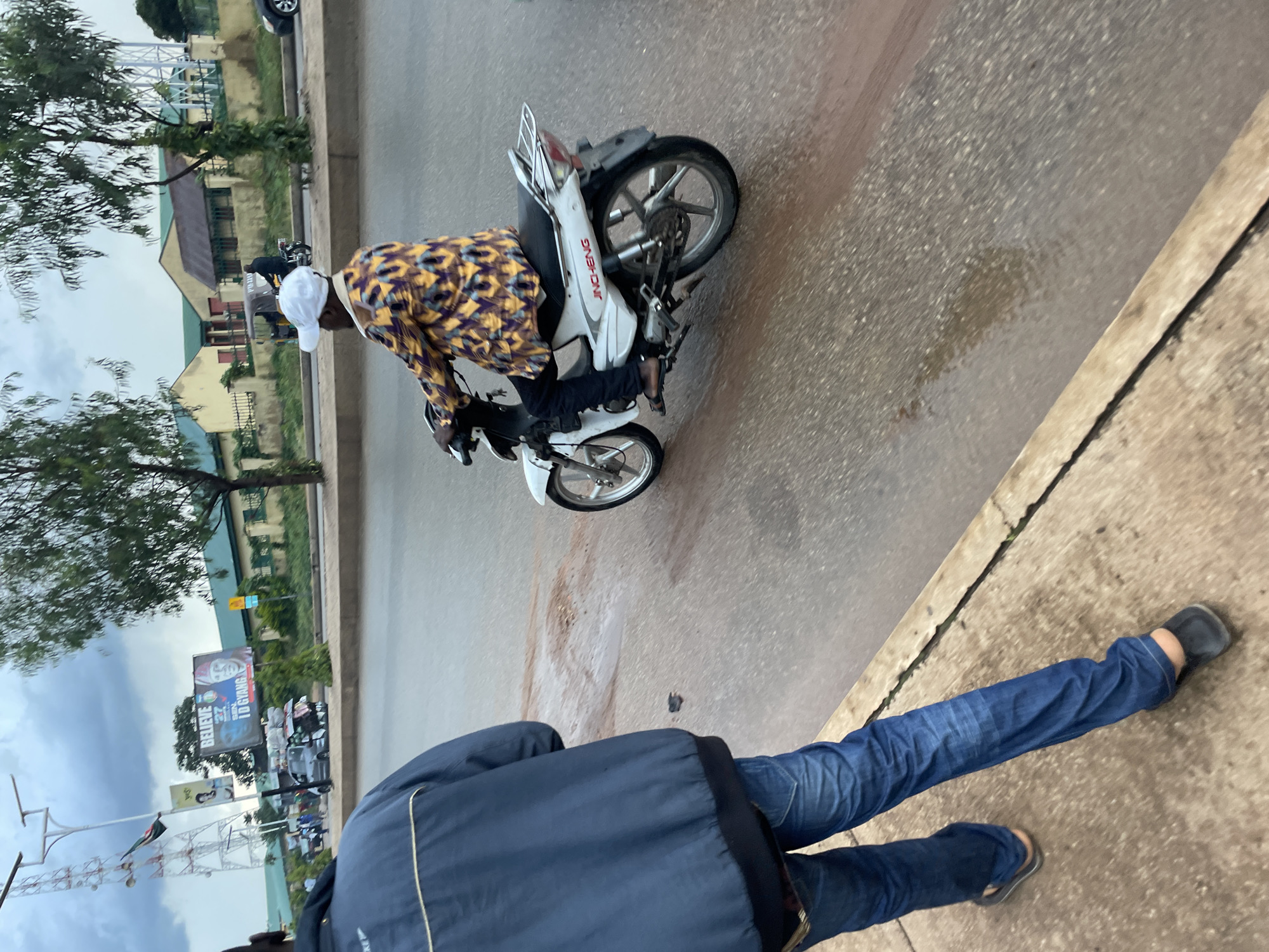 A man wearing a blue jacket and blue jeans walks on a sidewalk in front of a street in Jos, Nigeria. A man wearing a white cap and a yellow and purple patterned shirt rides a white motorcycle on the street. The street is wet, and there are trees and buildings in the background. A billboard with pictures of two men and the words "BELIEVE" and "TD GYANGB" is visible in the background. A communication tower with antennas is also visible on the left side of the image.