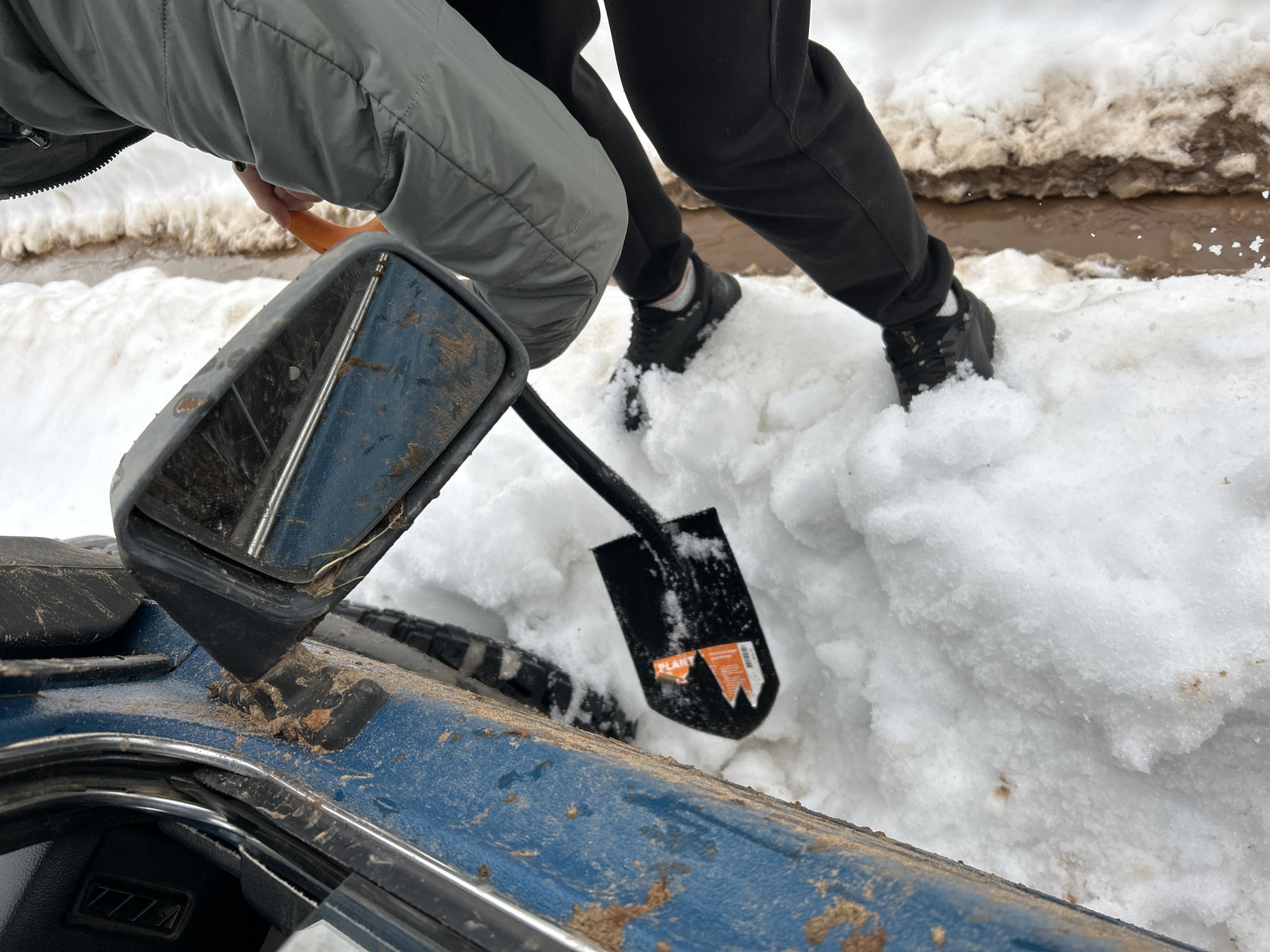 The image shows a blue vehicle stuck in the snow. Two people are visible near the vehicle. One person is wearing a grey jacket and holding an orange shovel handle. The other person is wearing black pants and shoes, standing in the snow. A black shovel is stuck in the snow near the vehicle's tire. The scene appears to be outdoors in a snowy environment. Based on the prompt, the location is assumed to be in or near Уральское, Russia. There is evidence of mud and dirty water where the snow is melting.