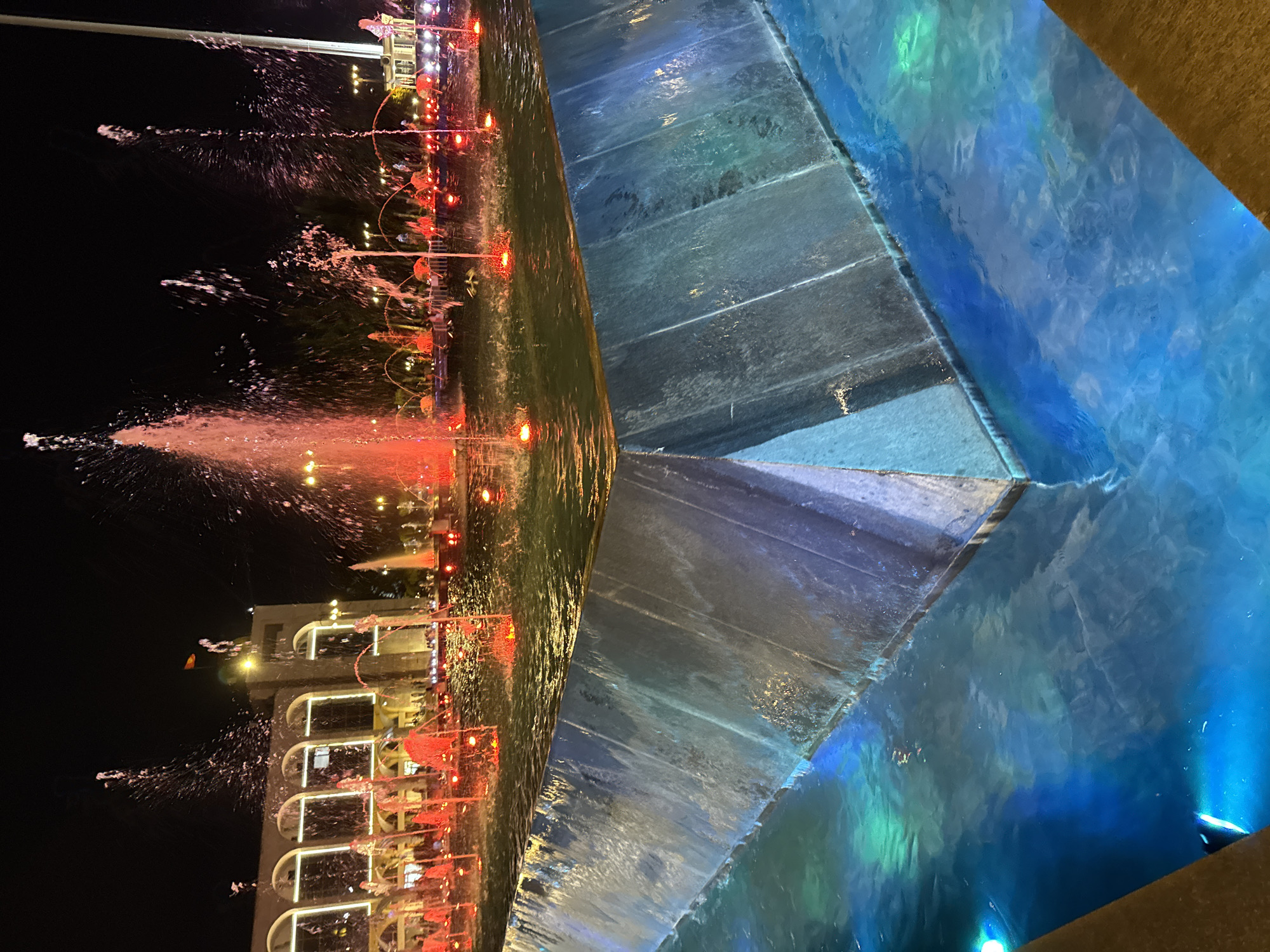An evening shot of the Chuy Avenue fountains in Bishkek, Kyrgyzstan, shows a large, multi-tiered fountain illuminated by red and blue lights. The foreground features a concrete structure of the fountain with water cascading down its sides. The middle ground displays several fountains with bright red lights at their base, shooting water into the night sky. In the background, an arched building is visible, with people milling about. The sky is dark, indicating it is nighttime.