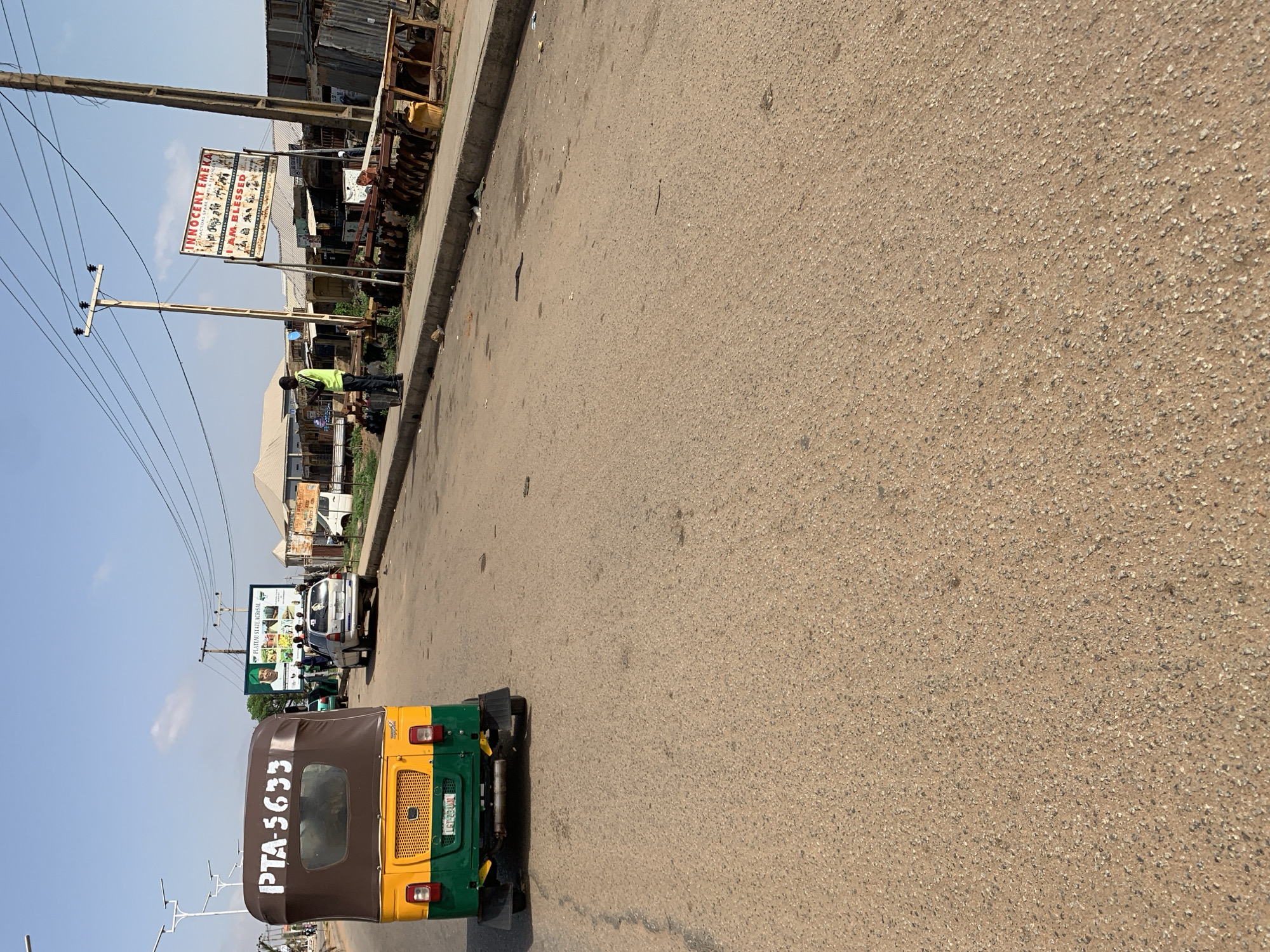 The image shows a street scene in Rafin Par, Nigeria. A brown, green, and yellow tricycle with the license plate "PTA-5633" is partially visible in the foreground. Further down the street, a white van is parked along the side. A billboard with images and text is visible behind the van. A person wearing a lime-green vest is standing near a storefront beneath a sign. Overhead, power lines stretch across the sky. The road is paved, and a curb separates it from the sidewalk, which is lined with various buildings.
