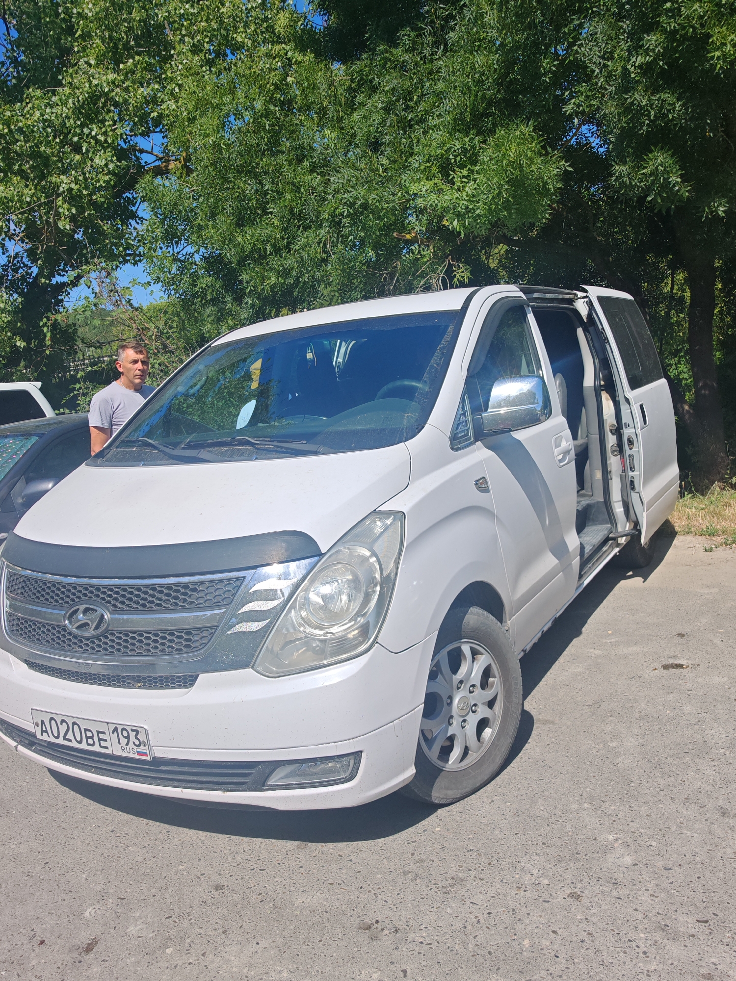 A man stands near a white Hyundai H-1 van parked on a paved road, with trees and foliage in the background. The van's front passenger door and sliding rear door are open. The license plate on the front of the van reads "A020BE 193 RUS". The man is wearing a grey t-shirt and appears to be looking towards the van. The scene is illuminated by daylight.