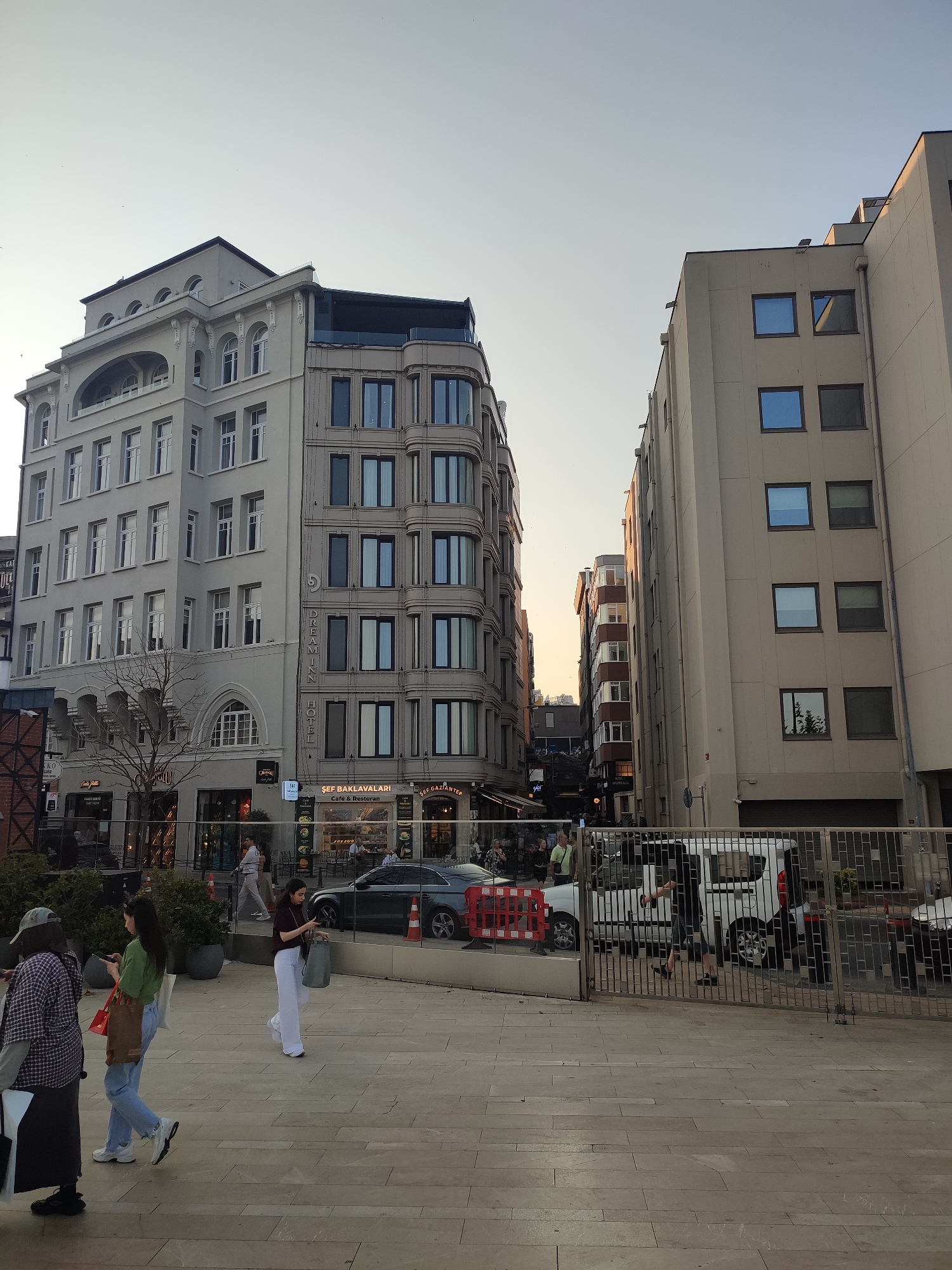 The image shows a street scene in the city of Beyoğlu, Turkey, characterized by modern architecture. On the left, a tall, light-colored building with numerous windows and decorative architectural details stands. Adjacent to it, on the right, is another tall building with a simpler, more modern facade featuring rows of blue-tinted windows. The sky above is clear and light, suggesting daytime. In the foreground, a paved walkway is visible, with several people walking. Some are carrying shopping bags. Behind them, a metal fence separates the walkway from a street where cars are parked and visible. Businesses are advertised on the buildings, including "Dream Inn Hotel" and "Şef Baklavaları Cafe & Restoran." The overall impression is of an urban environment with commercial activity.