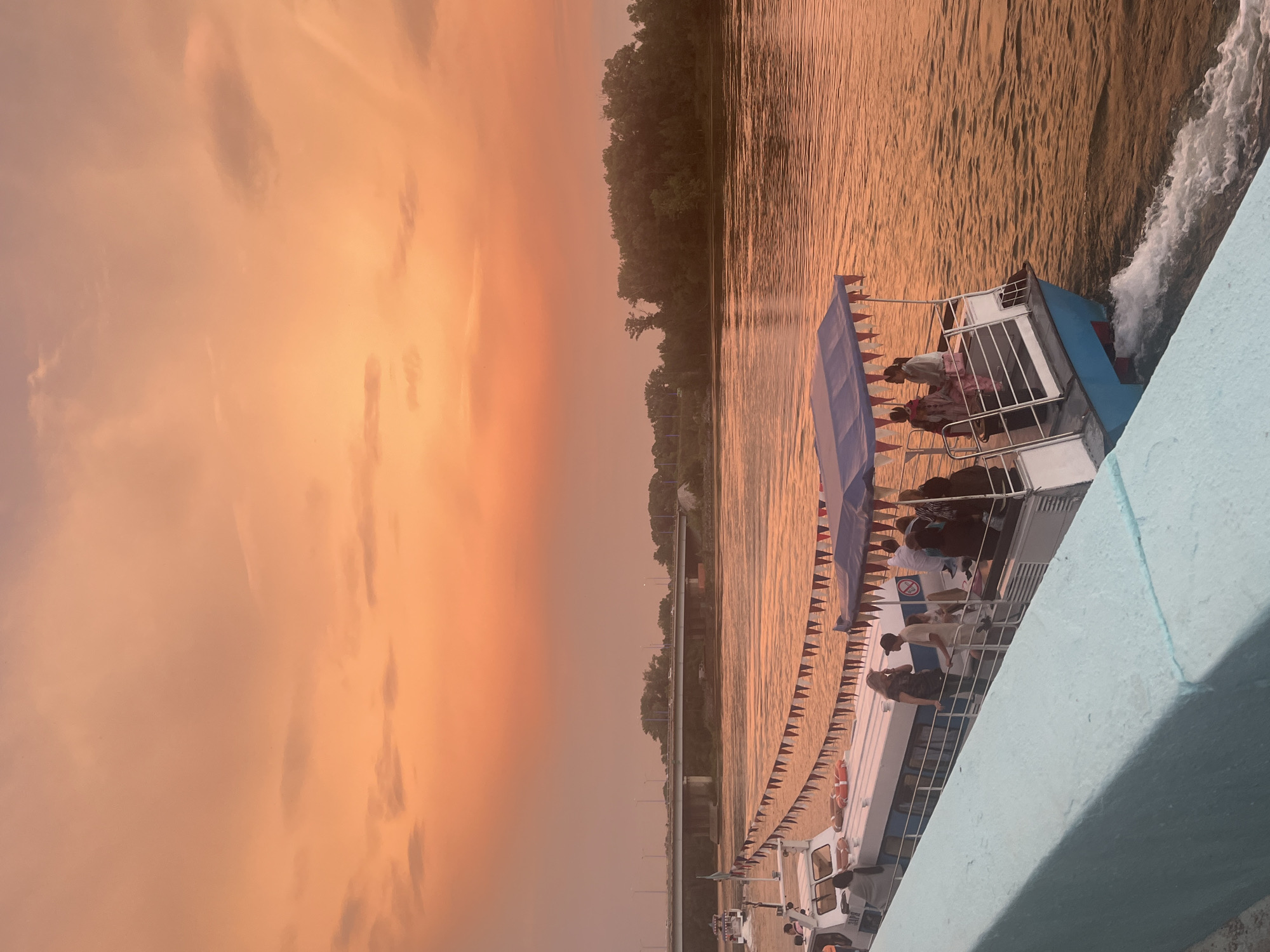 A view from a light blue colored barrier, looking out at a river during sunset. The sky is filled with an orange and pink gradient. A white tour boat, adorned with red and white pennant banners, is moving away from the viewer, creating a wake in the water. Several passengers are visible on the deck of the boat. In the background, a long bridge spans across the river, with trees lining the far bank. The overall impression is one of a peaceful evening scene in Oskemen, Kazakhstan.