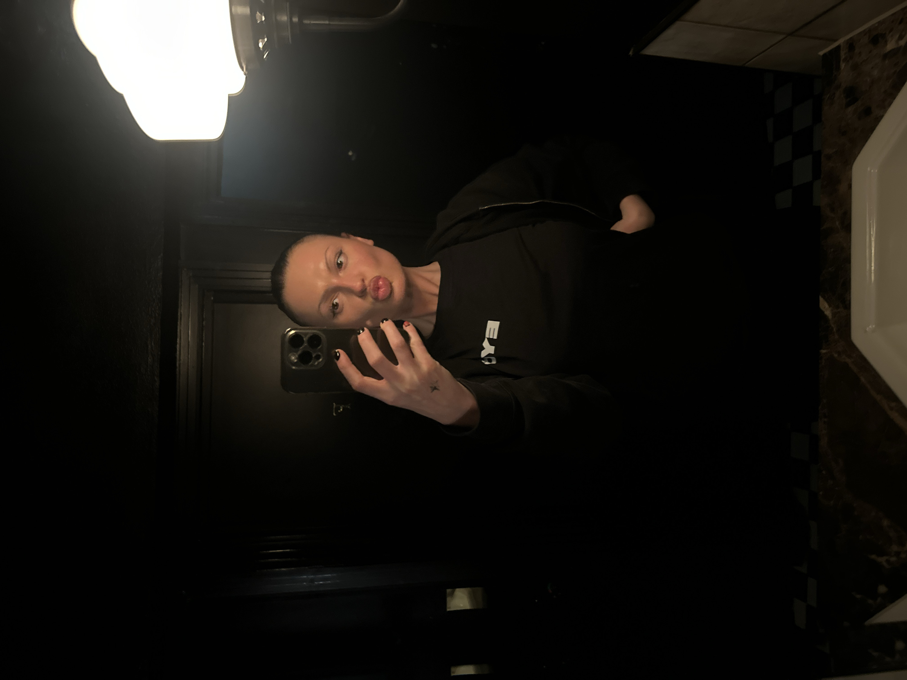 A person stands in a dark bathroom, taking a mirror selfie. They are wearing a black hoodie and have short hair. They are pouting at the camera while holding their phone in their right hand, which has black painted nails. A light fixture is visible above, and a sink is to the right. The bathroom has dark walls and a checkered pattern along the side of the sink.