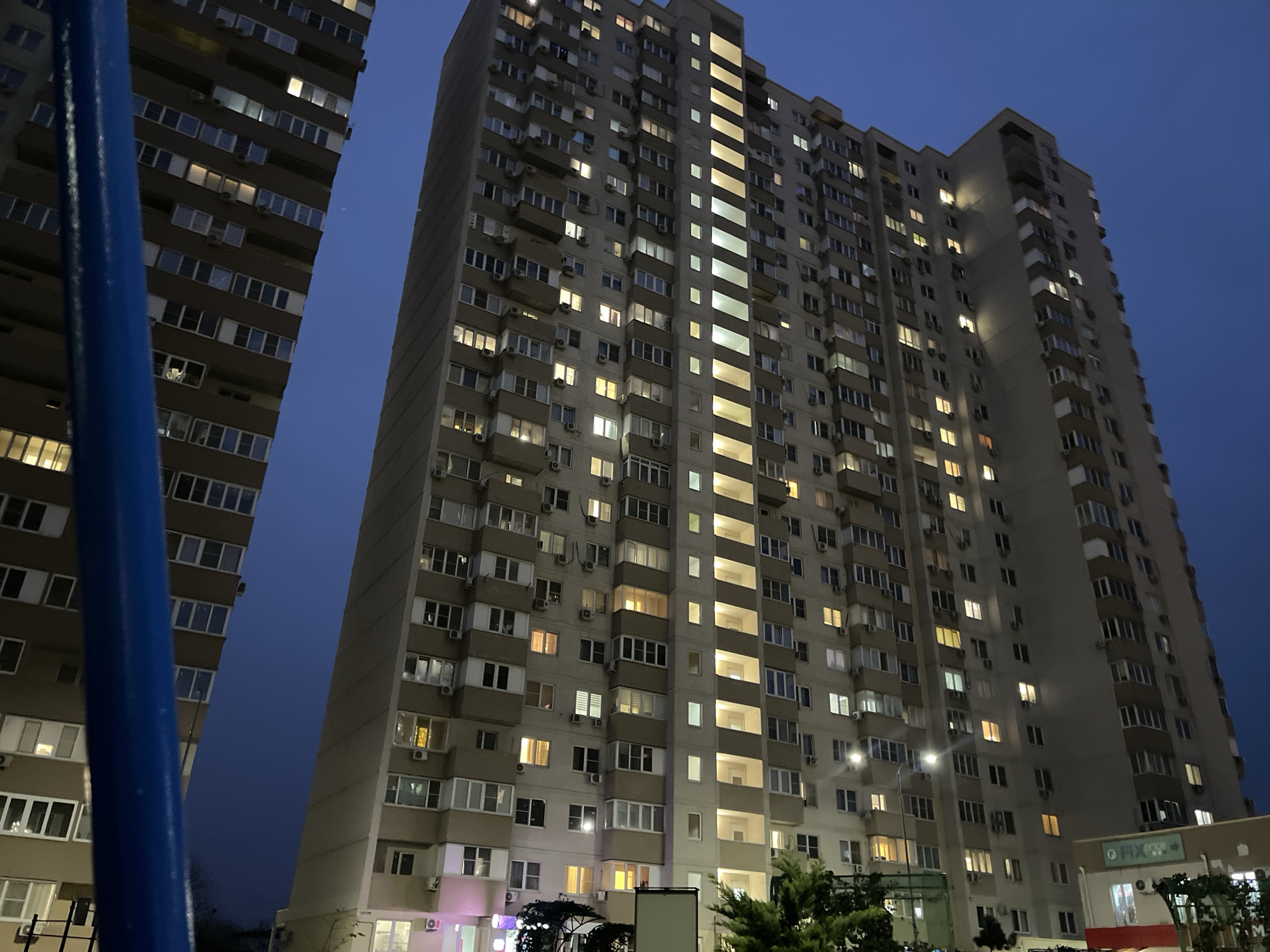 The image presents a nighttime or late twilight urban scene in Krasnodar, Russia, dominated by two tall, multi-story residential buildings set against a dark blue sky. In the left foreground, a prominent blue, curved structural element partially frames the view of the left building.
The central and largest subject is a beige high-rise apartment building on the right, which extends upwards out of the frame. Its facade is characterized by a repeating pattern of windows, many of which are brightly illuminated, casting a warm yellow glow. Numerous external air conditioning units are visible mounted below windows on almost every floor. The building also features recessed balconies and a distinctive vertical band of consistently lit windows or communal areas running down its center.
To the left, another tall apartment building, darker in tone, is partially visible, displaying similar architectural features and illuminated windows. The sky is a deep, clear blue, indicating either the late stages of dusk or the early hours of night.
At the base of the main building, on the lower right, glimpses of street-level activity are visible. A retail sign featuring the text "FIXPRICE" is clearly discernible in green lettering. There are also streetlights, faint purple lighting from possibly other storefronts, and dark green foliage from trees or bushes, adding to the urban setting. The overall atmosphere is calm and suggests the routine evening life of a busy residential area.
