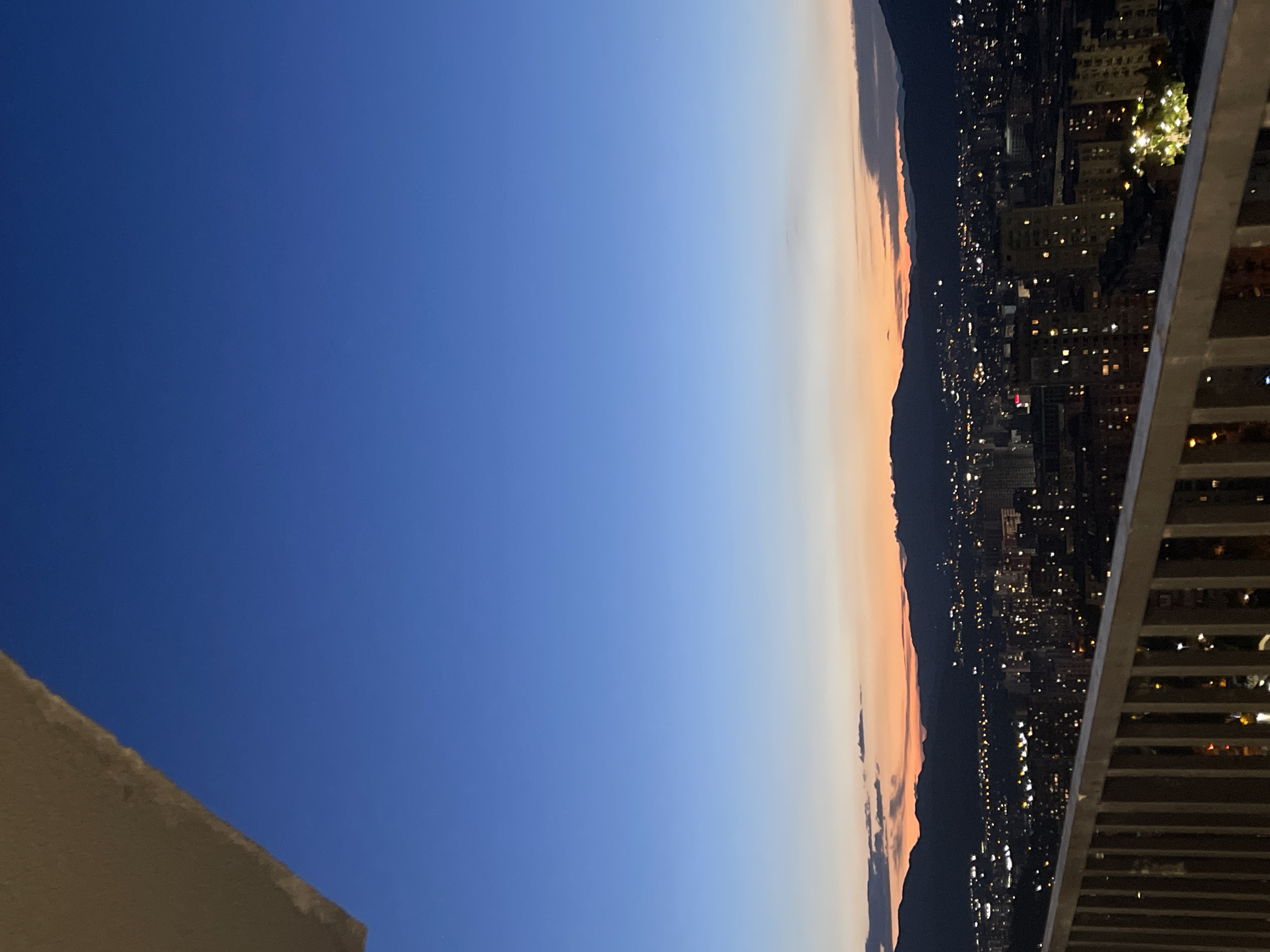 The image shows a cityscape of Tbilisi, Georgia, at dusk. A balcony railing is in the foreground, with a view of the city lights and mountains in the background. The sky transitions from a deep blue to an orange and pink hue as the sun sets. Buildings of various heights are visible, many with illuminated windows. The overall impression is one of a peaceful evening in the city.